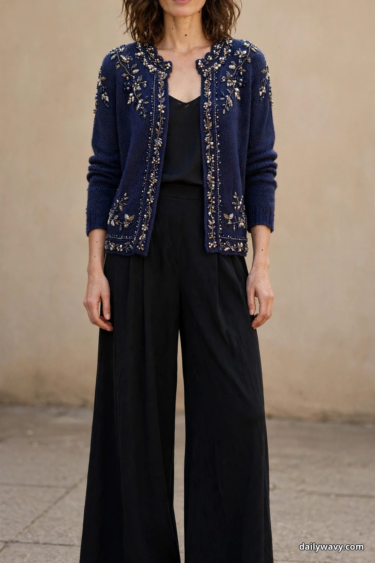 A woman stands against a beige wall in a chin-down frame wearing a navy beaded embellished cardigan open over a black camisole with black wide-leg crepe trousers that continue past the bottom edge of the frame