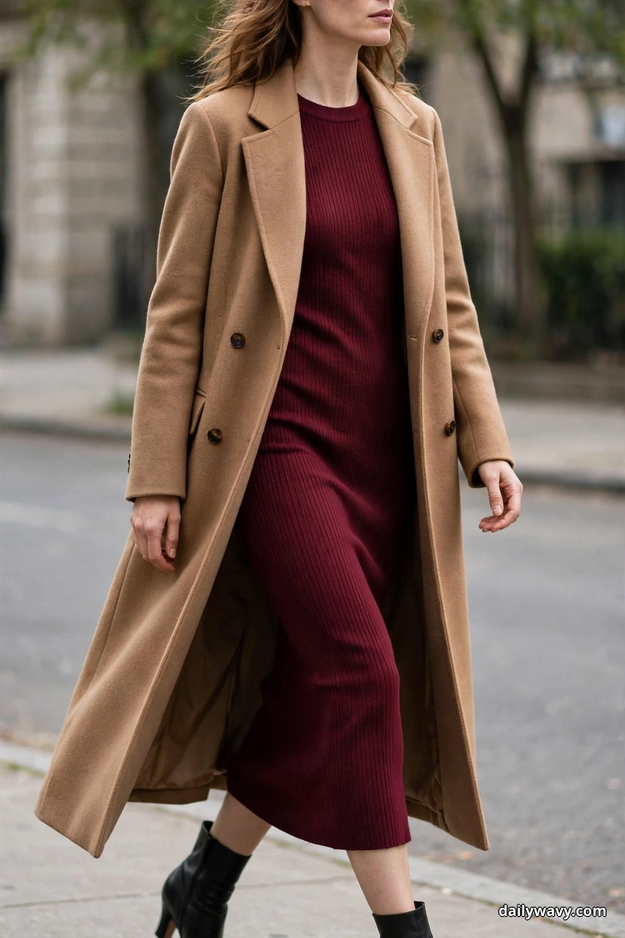 A woman walks along a city sidewalk with her face out of view wearing a burgundy ribbed knit midi dress under a long camel wool coat, with black heeled ankle boots