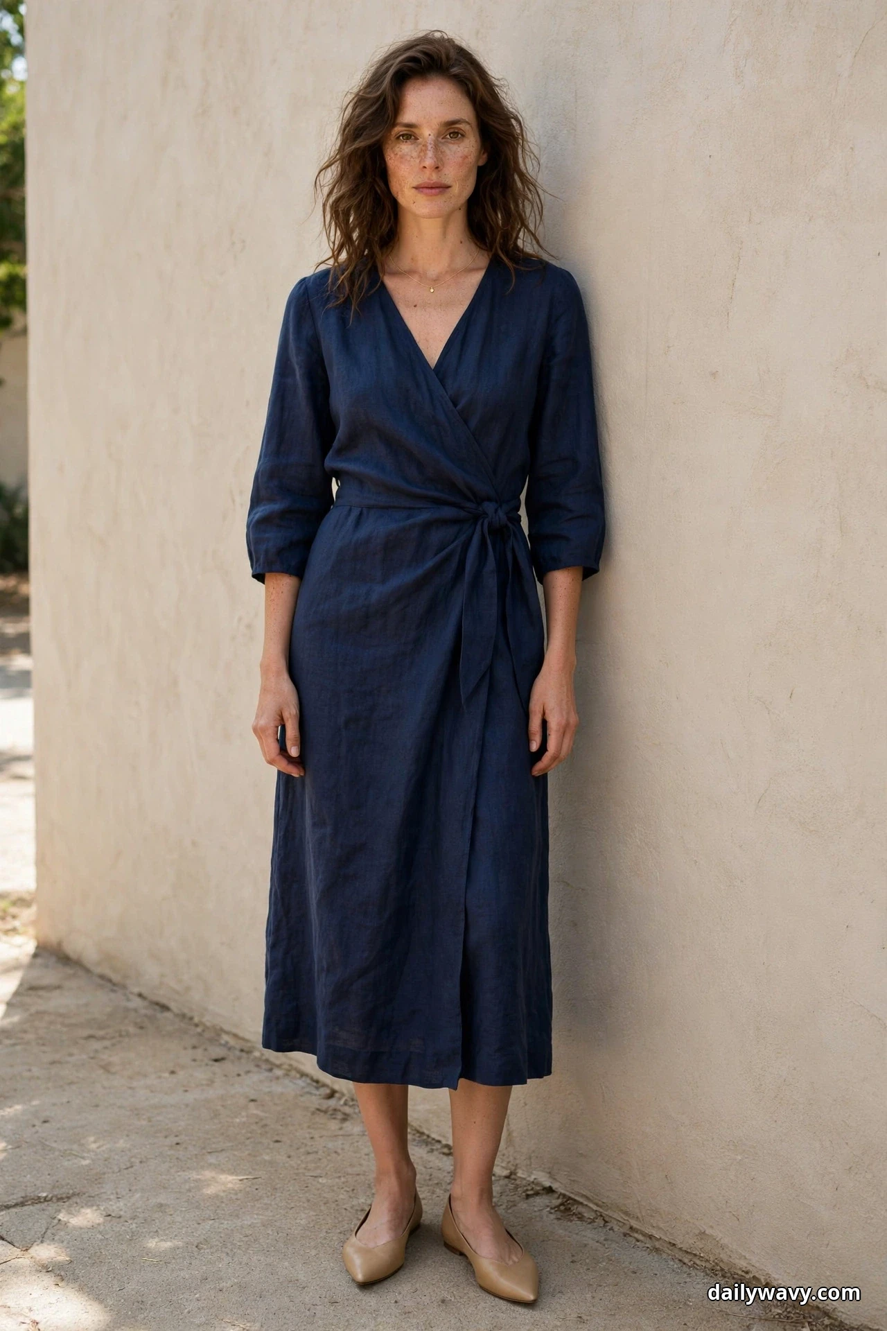 A woman stands full length against a plain wall wearing a navy wrap midi dress with a tied waist and three-quarter sleeves, a delicate necklace and nude pointed flats