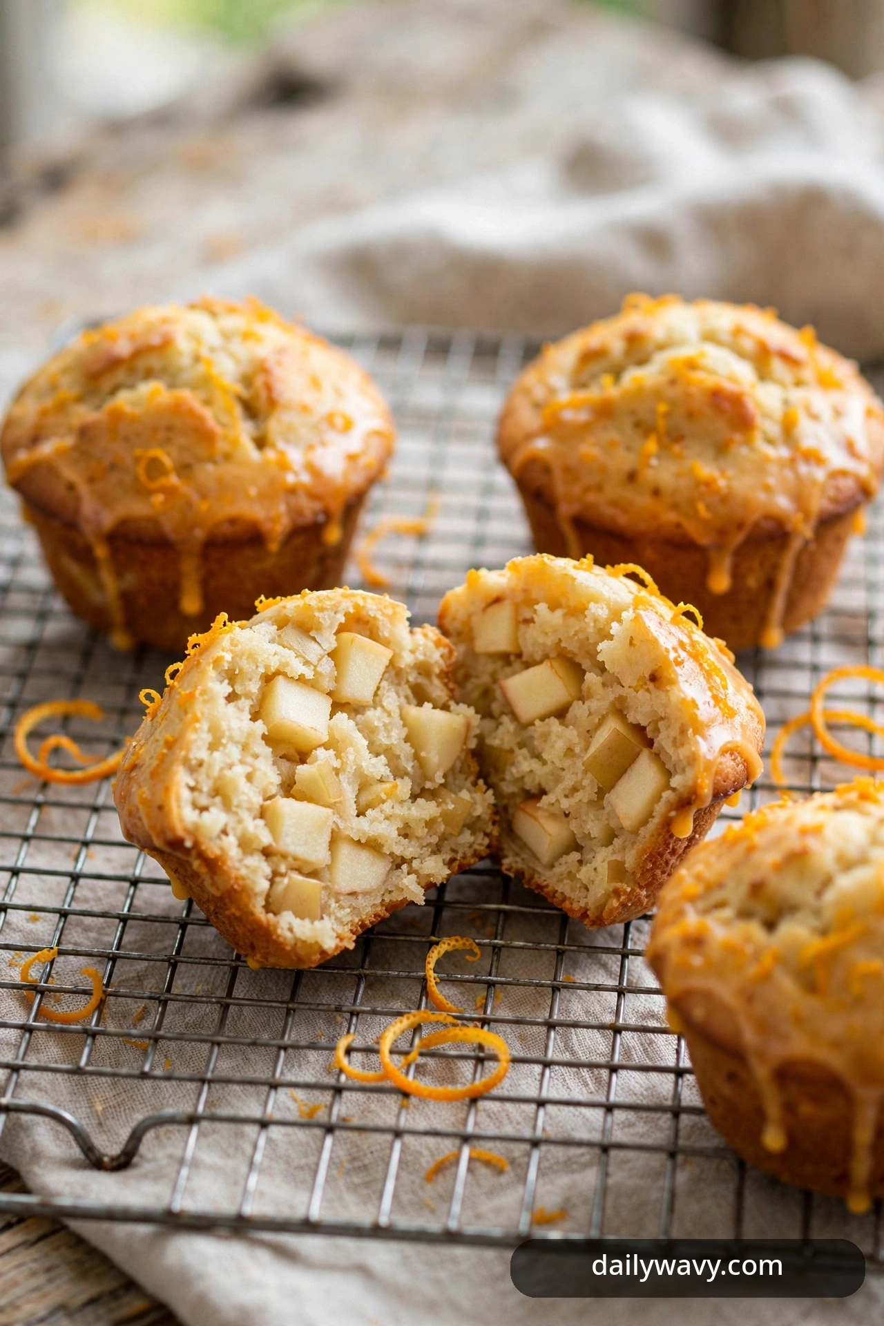 Orange-zest apple muffins with dripping glaze cool on a wire rack with orange peel curls scattered nearby, one split open to show the apple crumb.