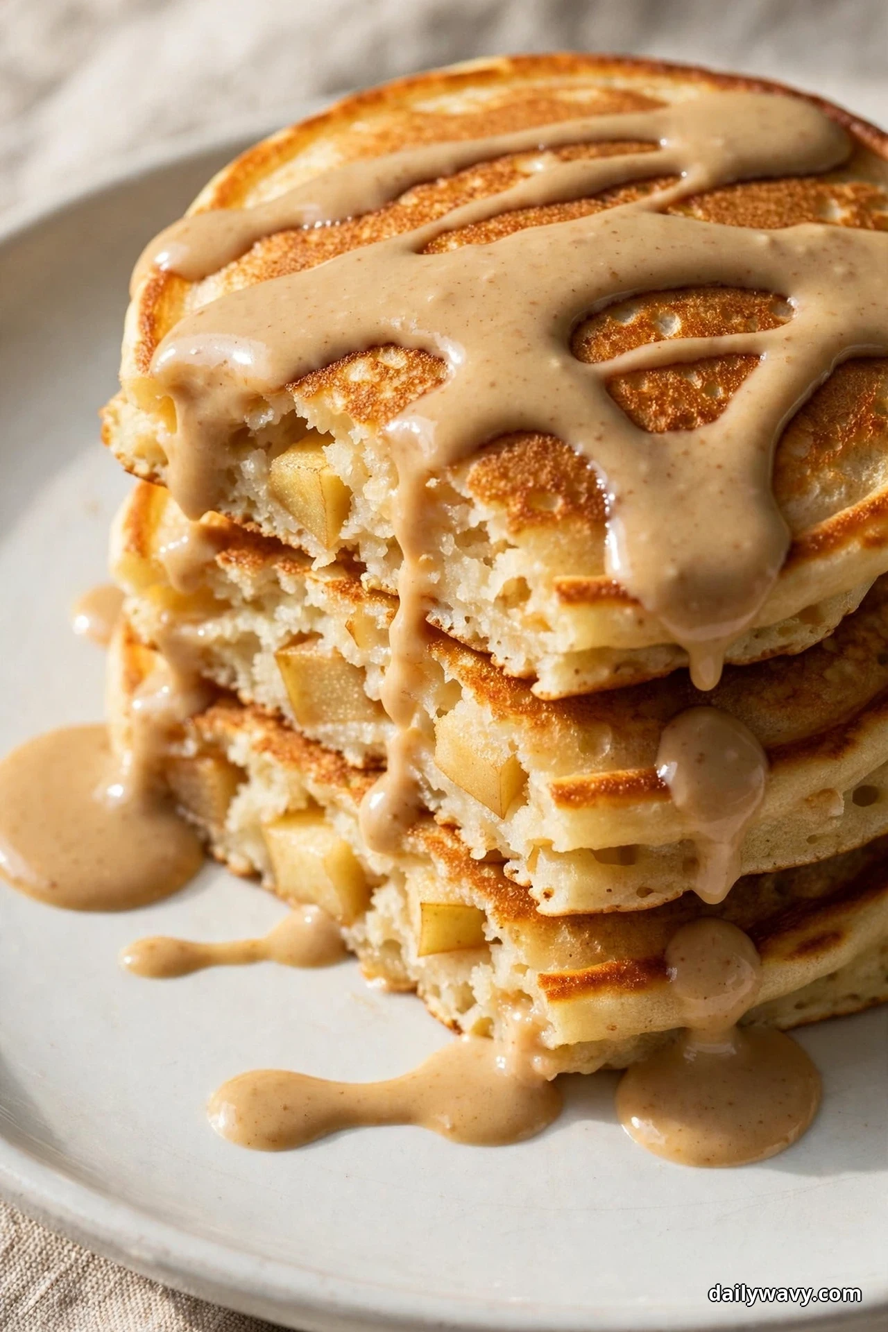 A close view of a stack of golden apple pancakes covered with thick tan tahini maple sauce dripping down the sides and pooling on a white plate, with apple chunks visible in the cut faces