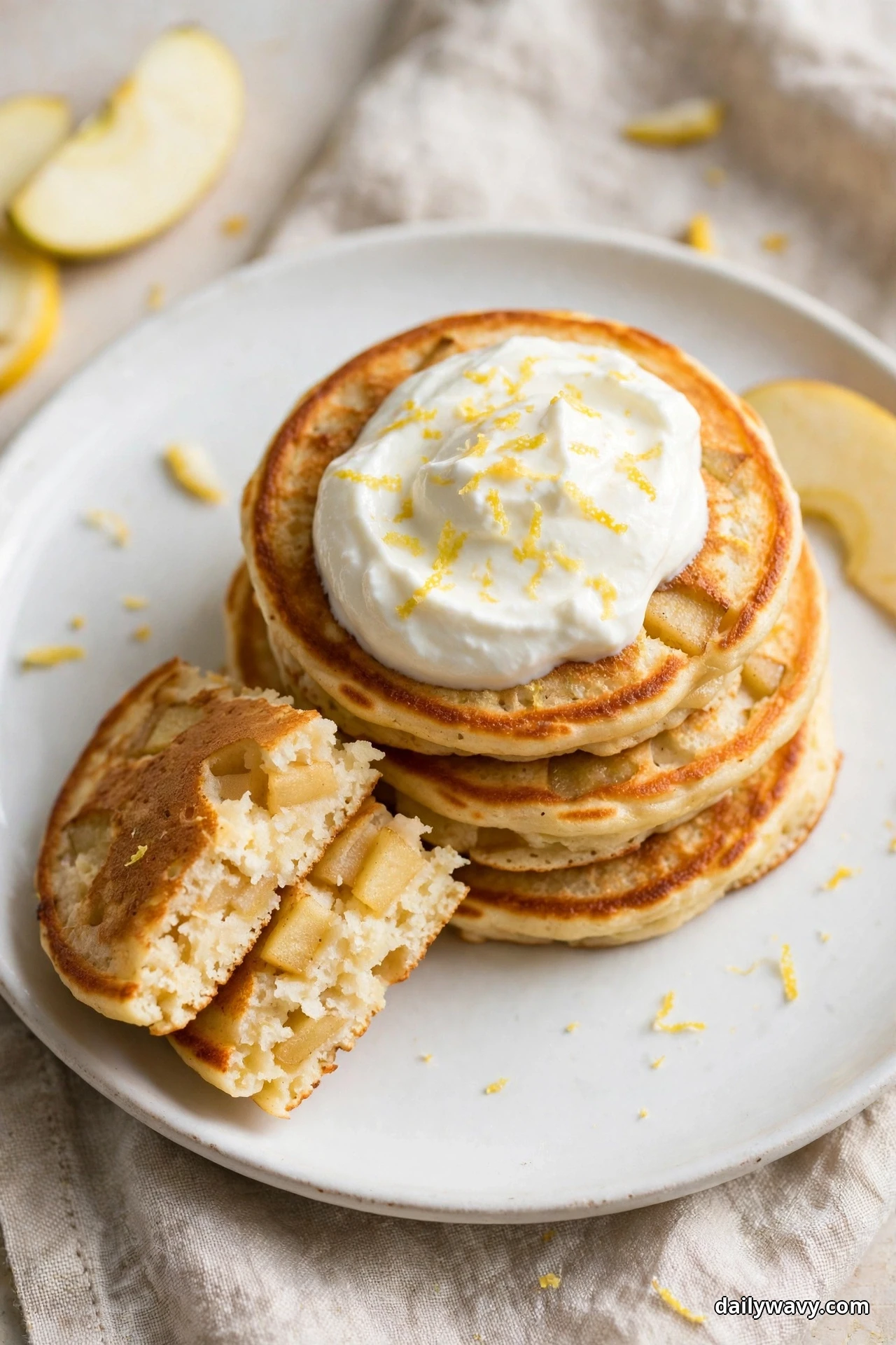 A stack of golden apple pancakes topped with a large dollop of white yogurt sprinkled with lemon zest strands sits on a white plate, with one pancake pulled aside and broken open showing the apple chunks inside