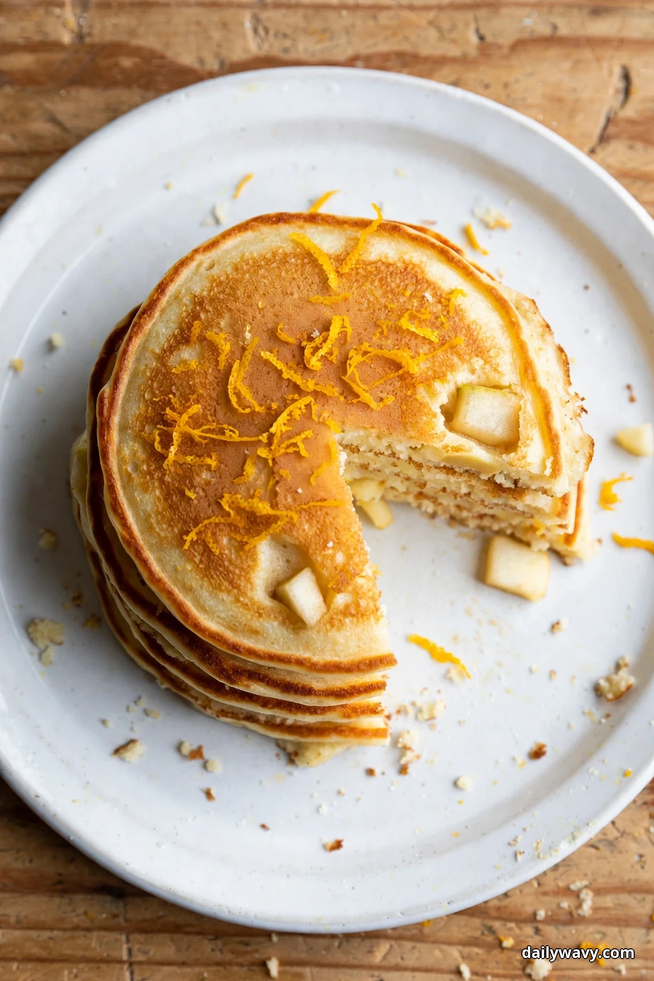 Seen from above, a stack of golden apple pancakes sprinkled with fine orange zest strands sits on a white plate, one wedge cut out showing the layered crumb and apple cubes