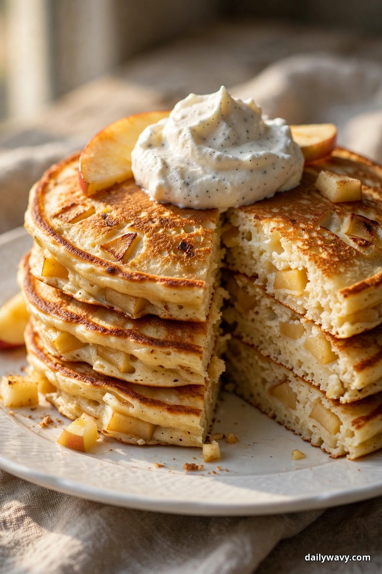 A tall stack of golden apple pancakes topped with a dollop of whipped cream speckled with vanilla bean seeds sits on a white plate, one wedge cut away showing the apple chunks inside the fluffy crumb