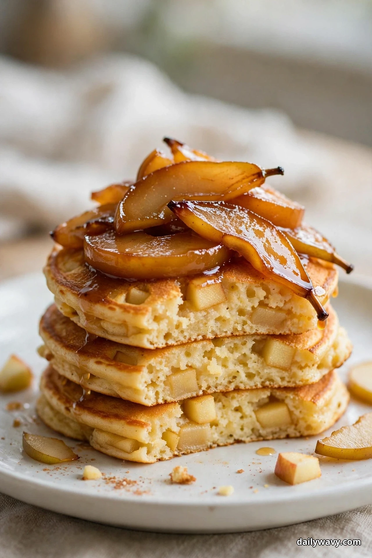 A stack of golden apple pancakes topped with glossy caramelized pear slices sits on a white plate, the open front faces showing the apple chunks baked into the fluffy crumb