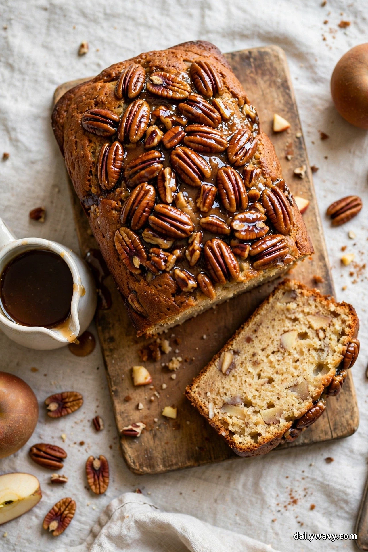 Seen from above, an apple quick bread loaf topped with glossy maple-glazed pecan halves rests on a wooden board, with one slice cut in front and a small jug of maple syrup beside it