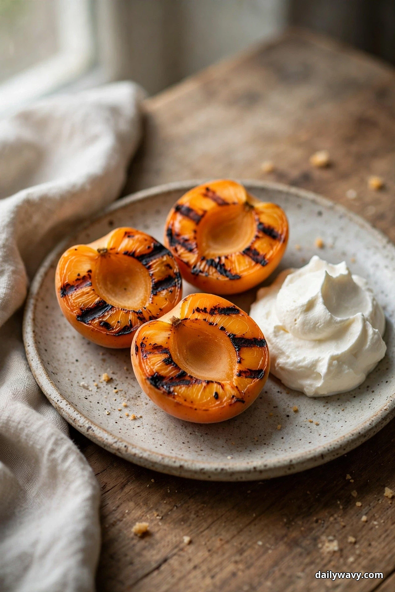 Three grilled apricot halves with dark char marks sit on a speckled plate beside a dollop of whipped cream