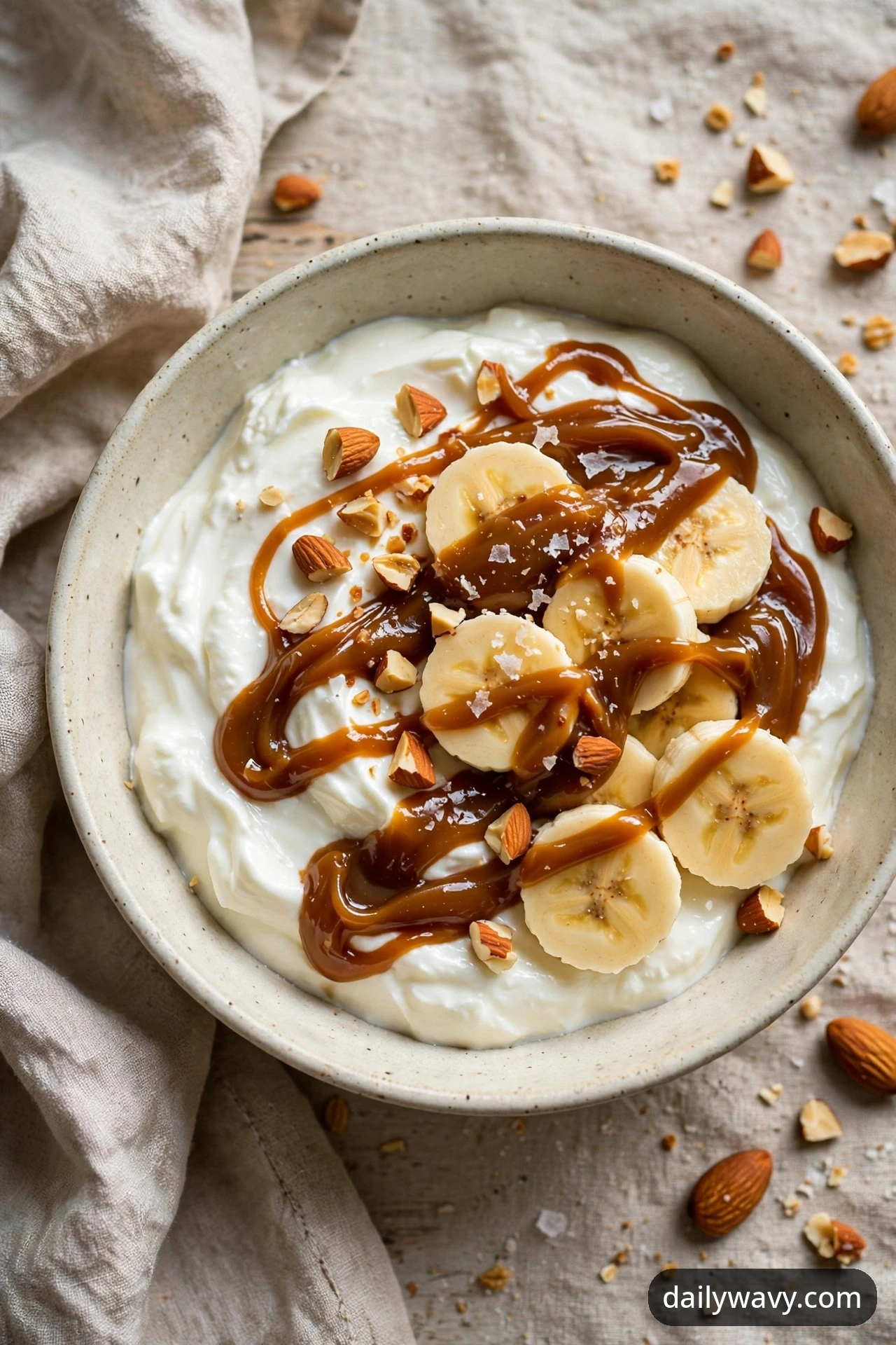 An overhead view of a banana yogurt bowl with date caramel ribbons, banana coins, chopped toasted almonds and flaky salt over yogurt, with whole almonds scattered on a linen cloth beside the bowl.