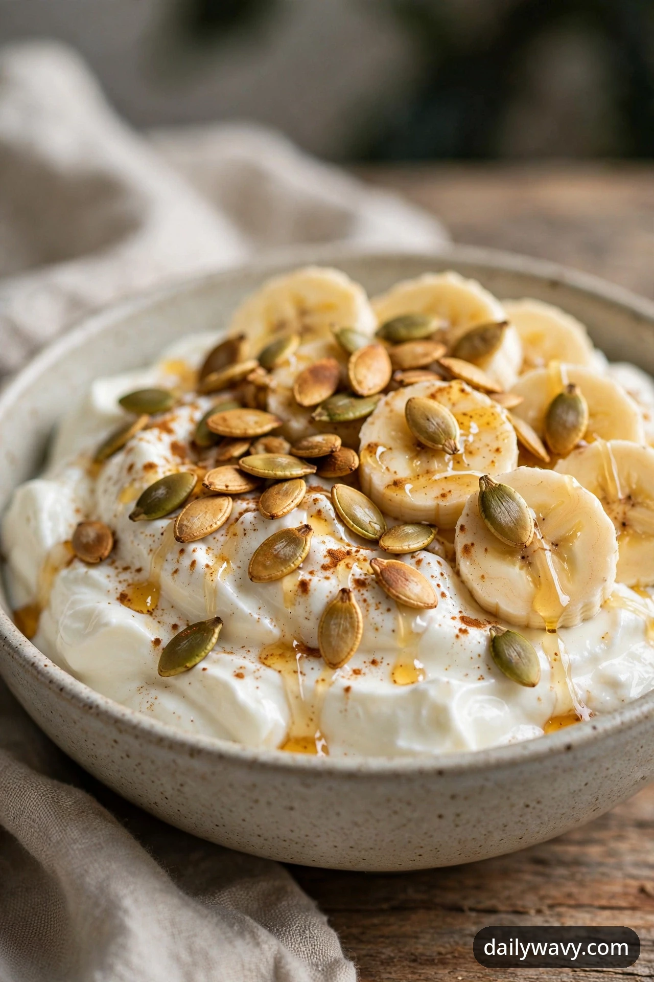 A banana yogurt bowl with toasted pumpkin seeds, a honey drizzle and a cinnamon dusting over yogurt and banana coins, photographed close on a wooden table.