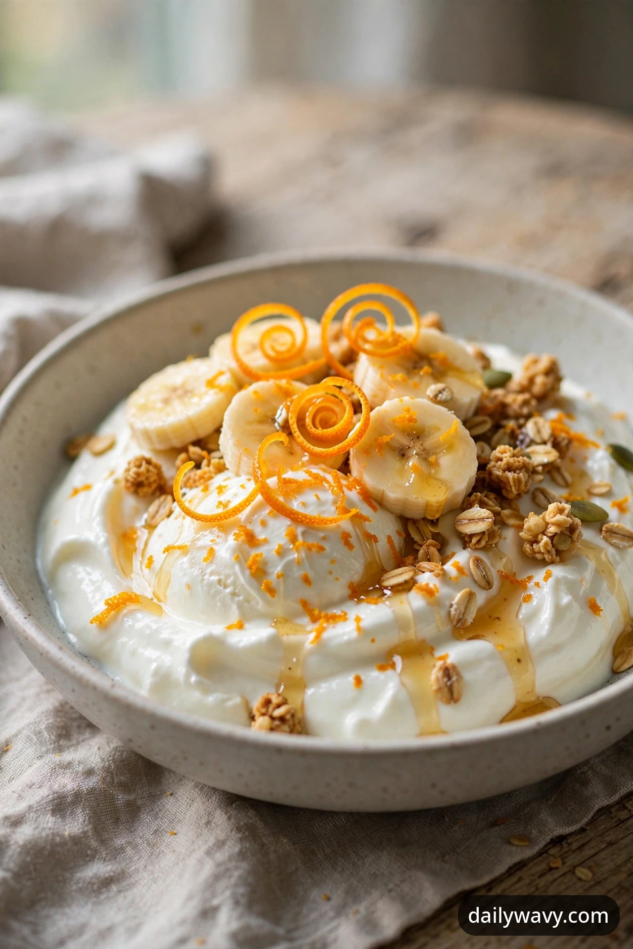A banana yogurt bowl with curled orange zest spirals, fine orange zest, granola and a honey drizzle over yogurt and banana coins, photographed on a linen napkin.