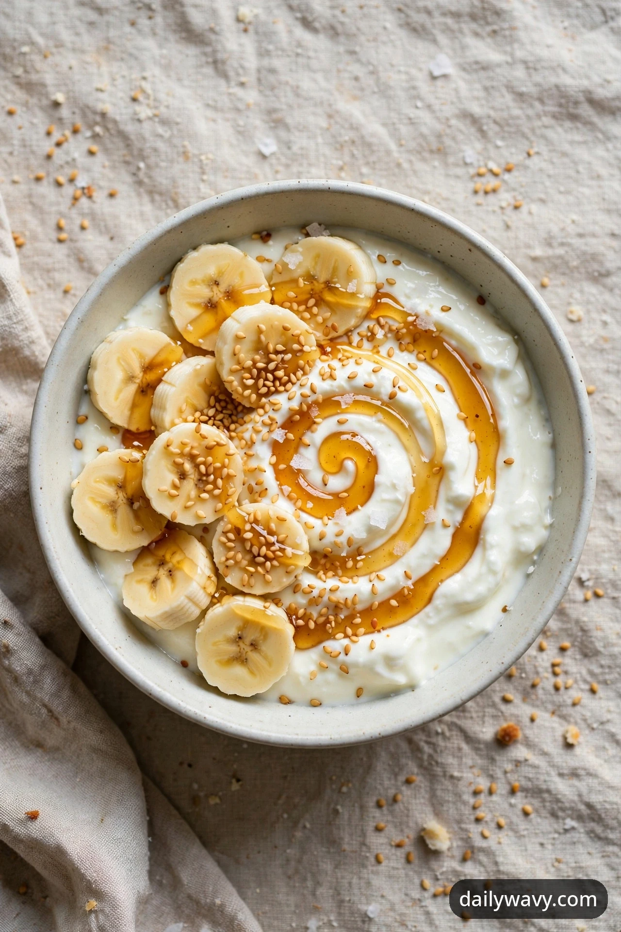 An overhead view of a banana yogurt bowl with honey drizzled in a spiral, toasted sesame seeds and flaky salt over yogurt and banana coins on a linen cloth.