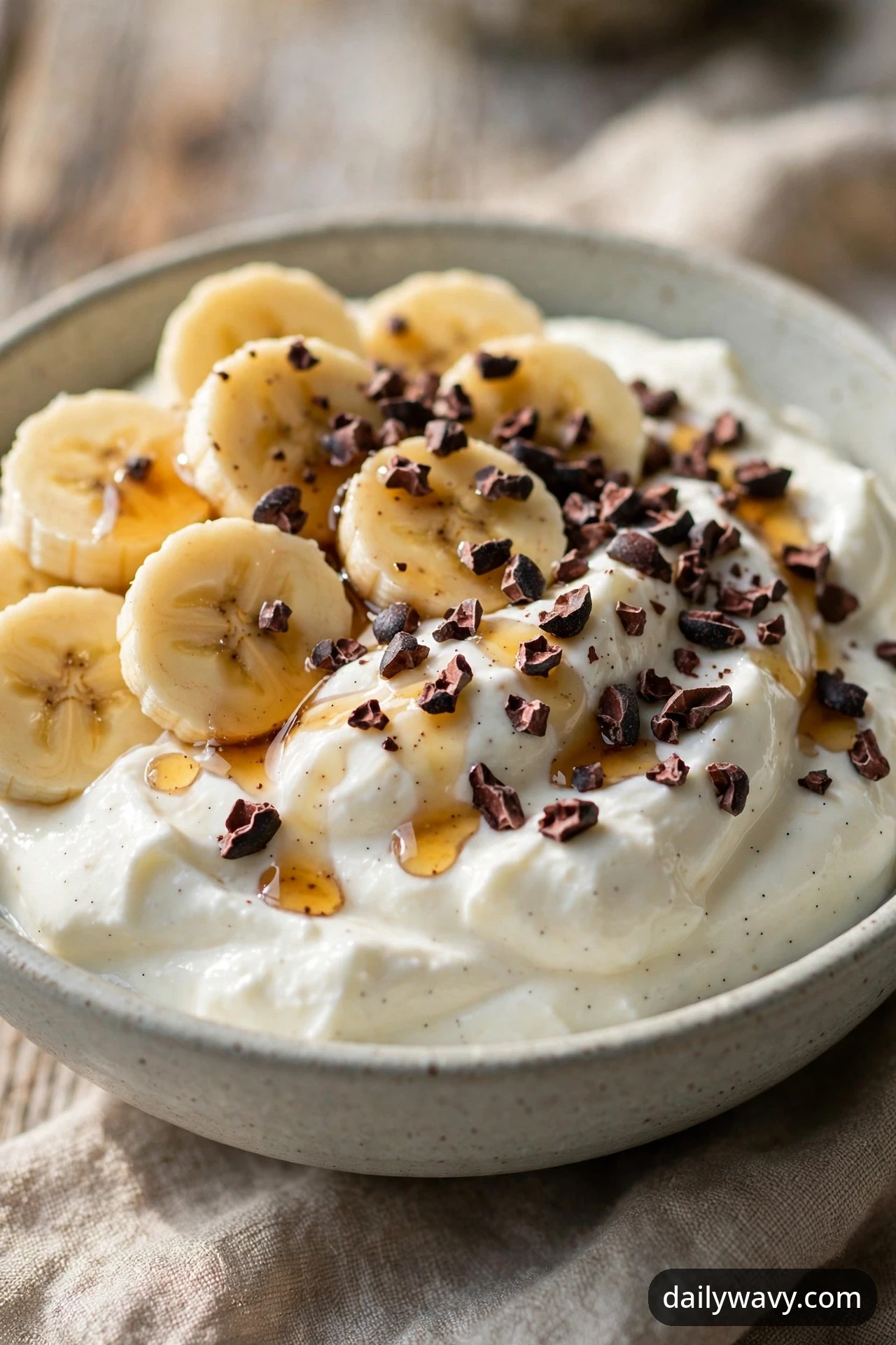 A banana yogurt bowl with cocoa nibs and a maple drizzle over yogurt and banana coins, photographed close on a wooden table.