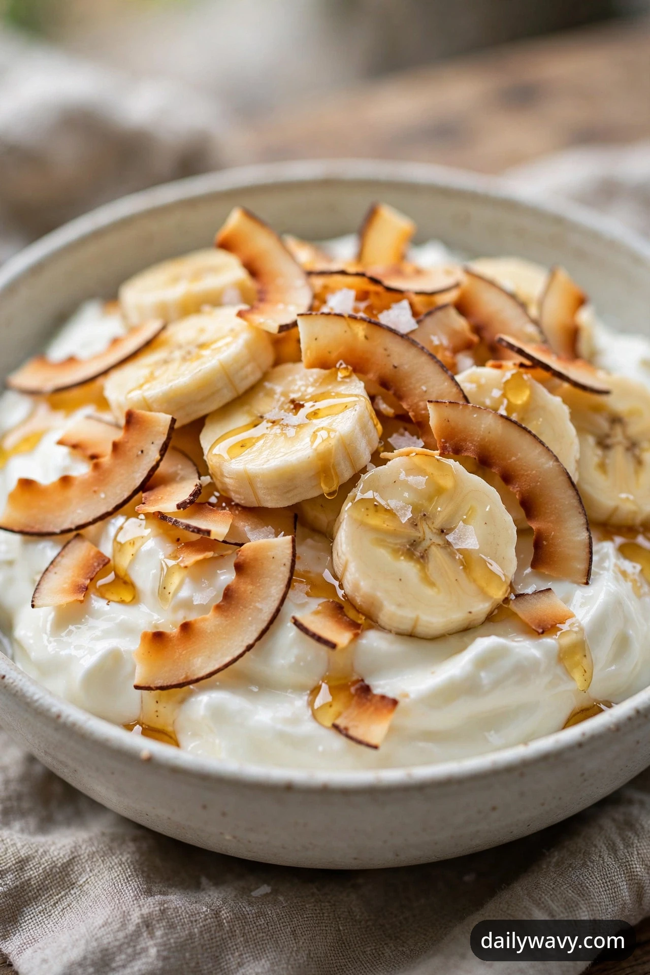 A banana yogurt bowl with wide toasted coconut flakes, flaky salt and a honey drizzle over yogurt and banana coins, photographed close.