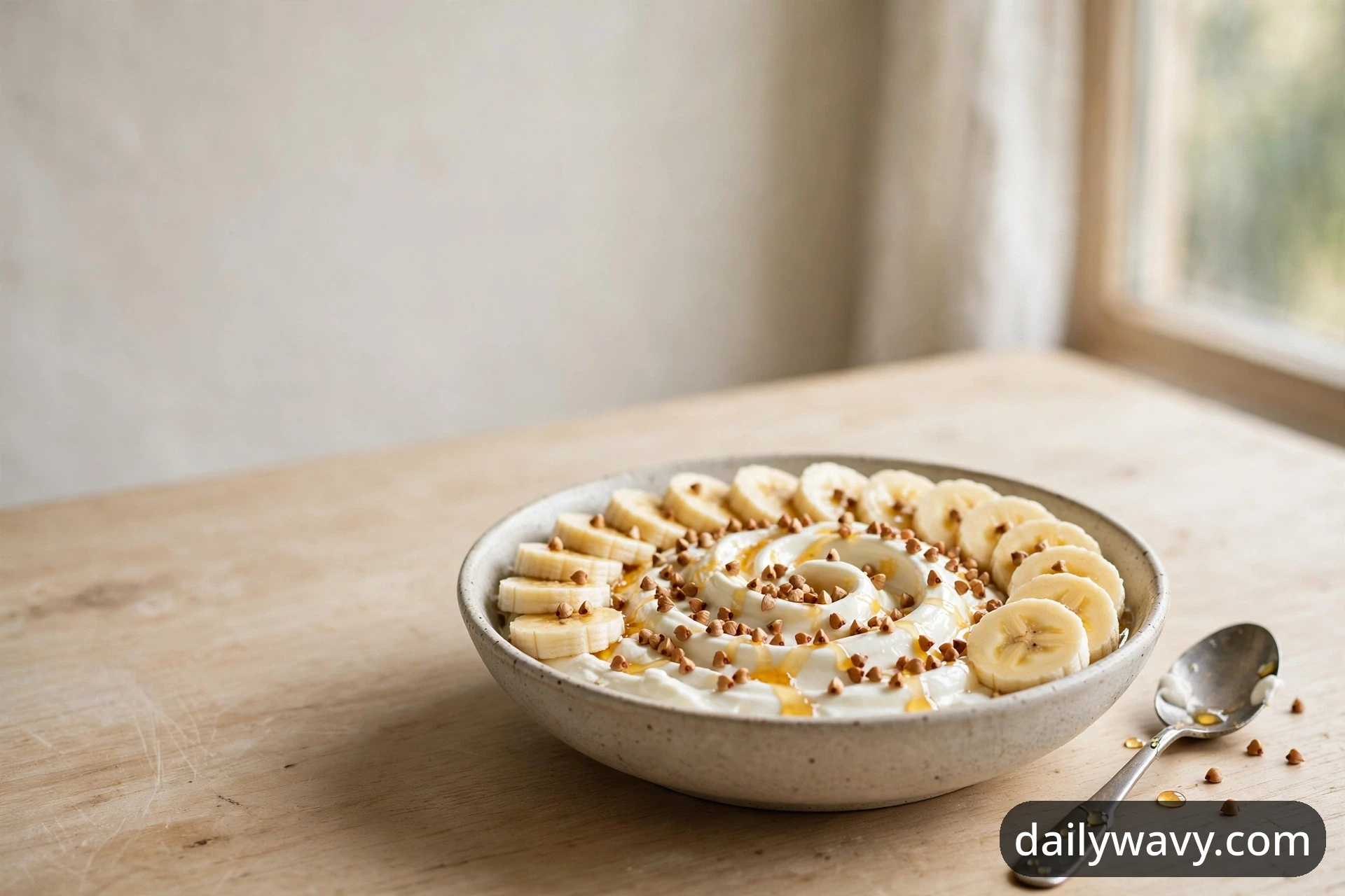 A wide view of a banana yogurt bowl with buckwheat crunch, swirled yogurt, a ring of banana slices and a honey drizzle in a shallow ceramic bowl on a wooden table by a window, with a spoon beside it.
