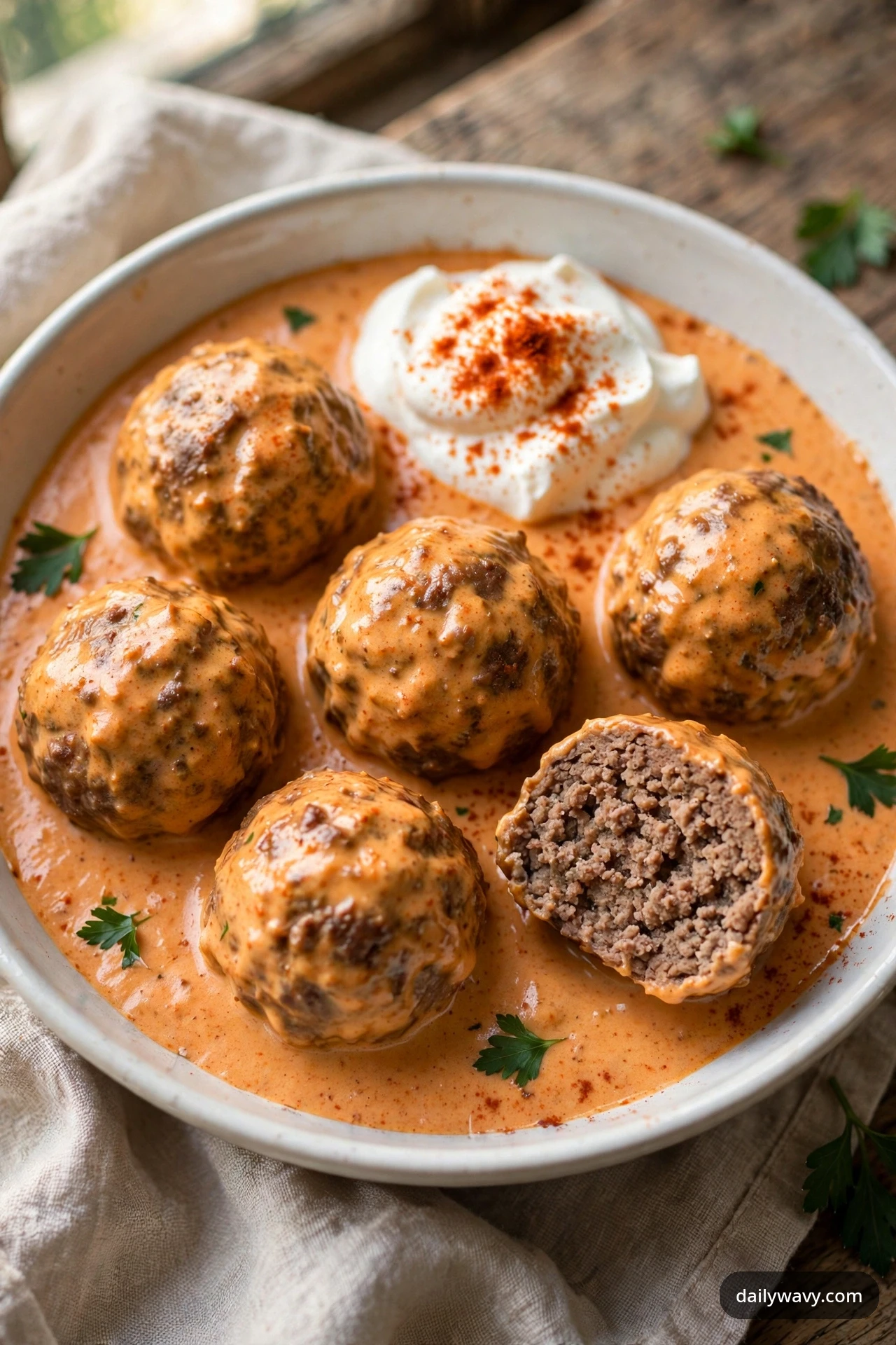 Beef meatballs in creamy paprika-orange sauce sit in a white bowl, one cut open to reveal the cooked interior, with a dollop of sour cream dusted with paprika and scattered parsley.