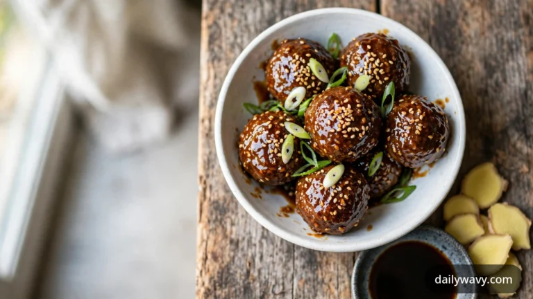 Glossy beef meatballs in sesame ginger glaze are piled in a shallow white bowl, topped with toasted sesame seeds and scallion curls, with fresh ginger slices and a small dish of dark dipping sauce on the wooden table beside it.
