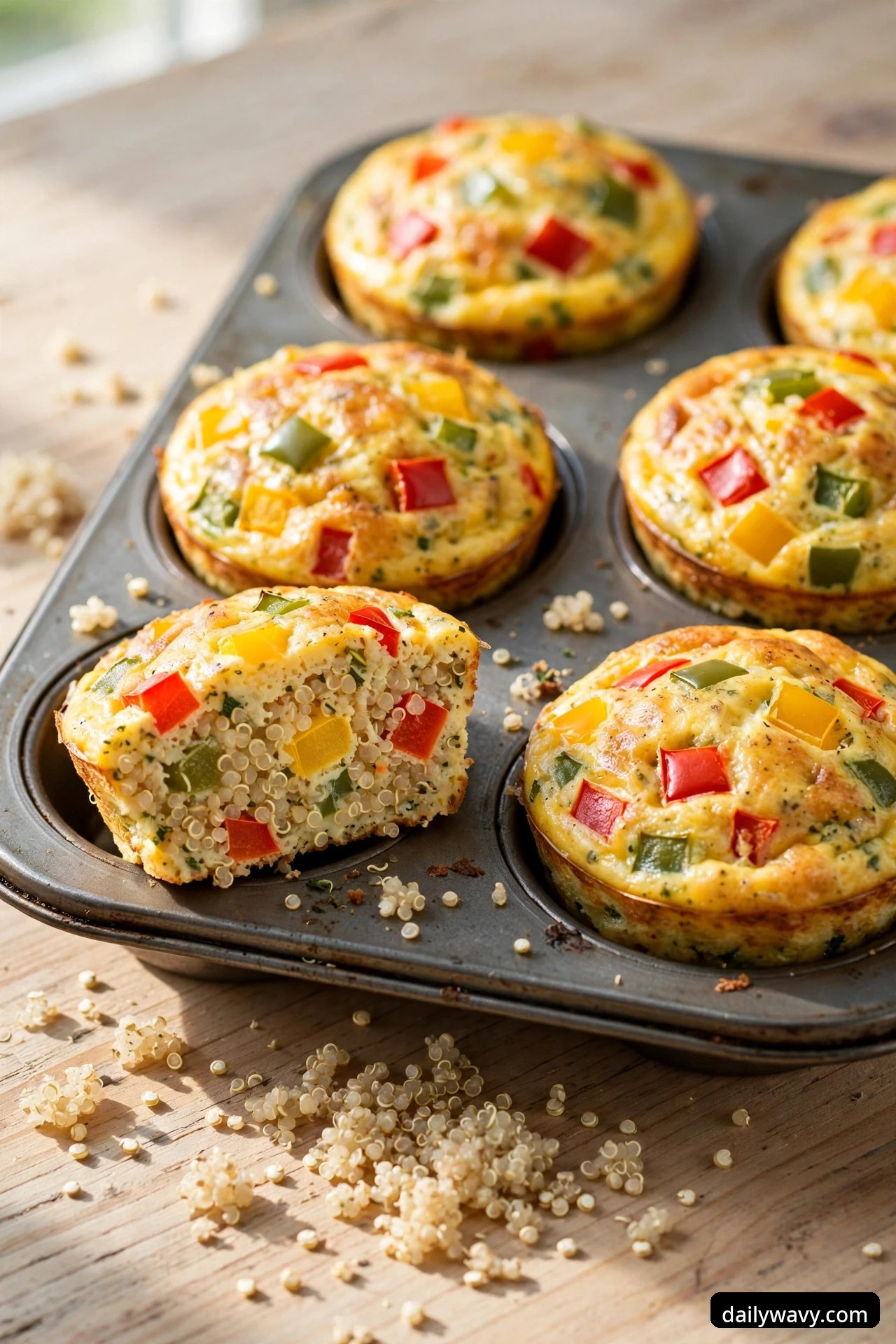 Bell pepper egg muffins sit in a muffin tin with one muffin cut open at the front showing cooked quinoa grains and red, yellow, and green pepper dice throughout the set egg