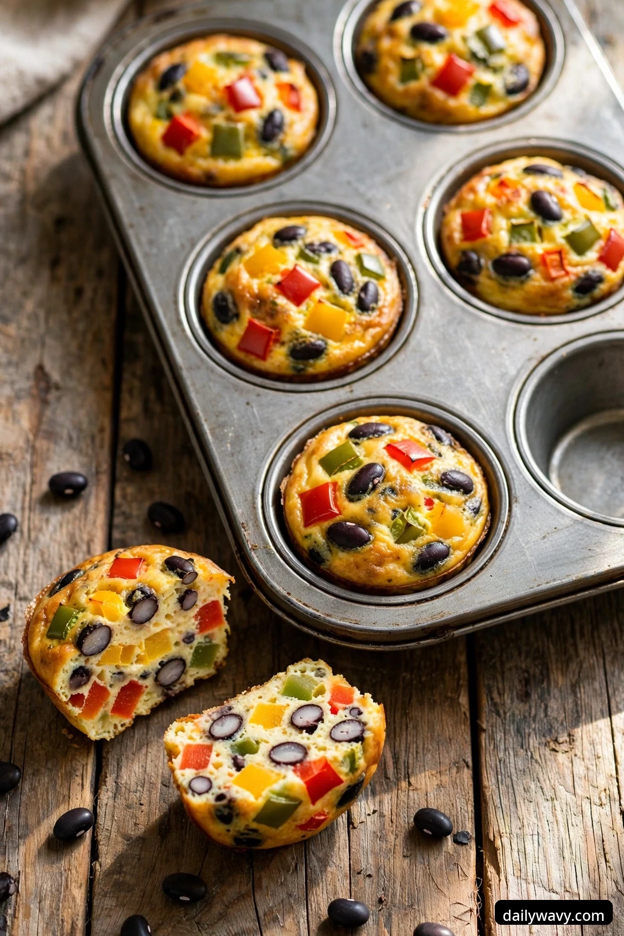 Bell pepper egg muffins with whole black beans baked into the tops sit in a muffin tin, with two halves on the wooden table showing black beans and pepper dice inside
