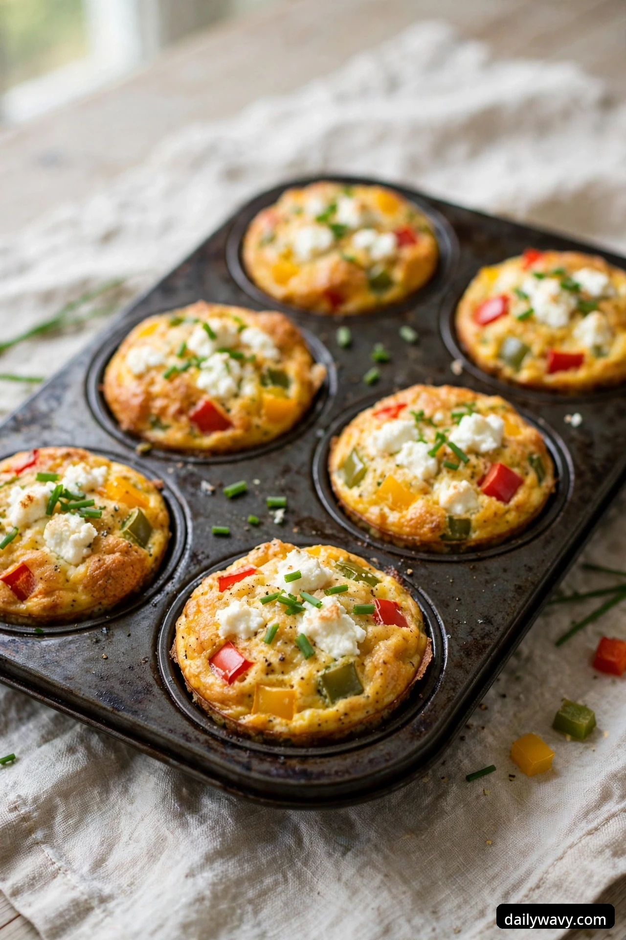 Bell pepper egg muffins topped with white goat cheese crumbles and snipped chives sit in a muffin tin, with red, yellow, and green pepper dice visible in the golden egg