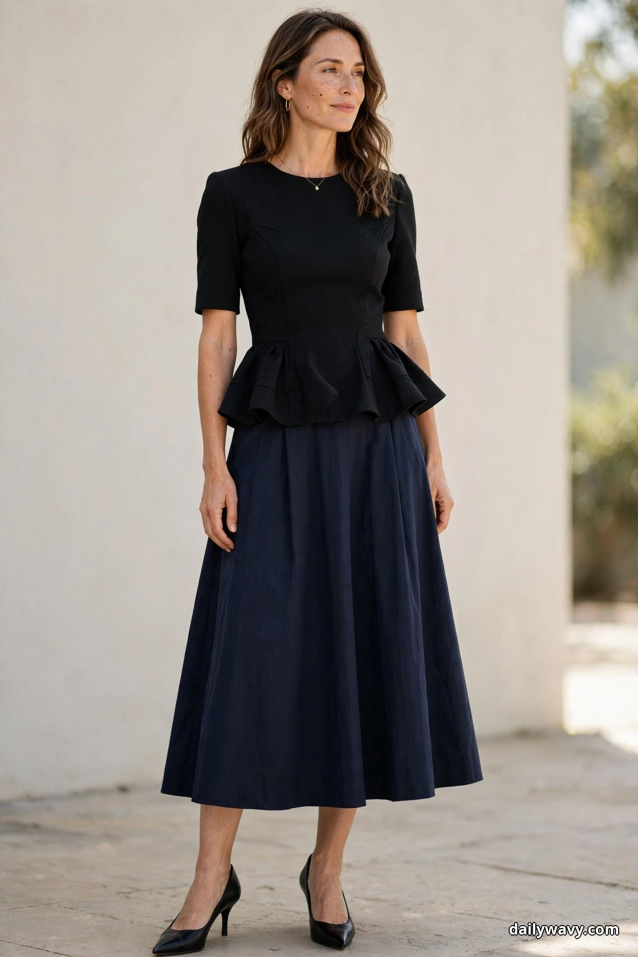 A woman stands full length outdoors against a plain cream wall wearing a black short-sleeve peplum top with a navy A-line midi skirt and black pointed pumps