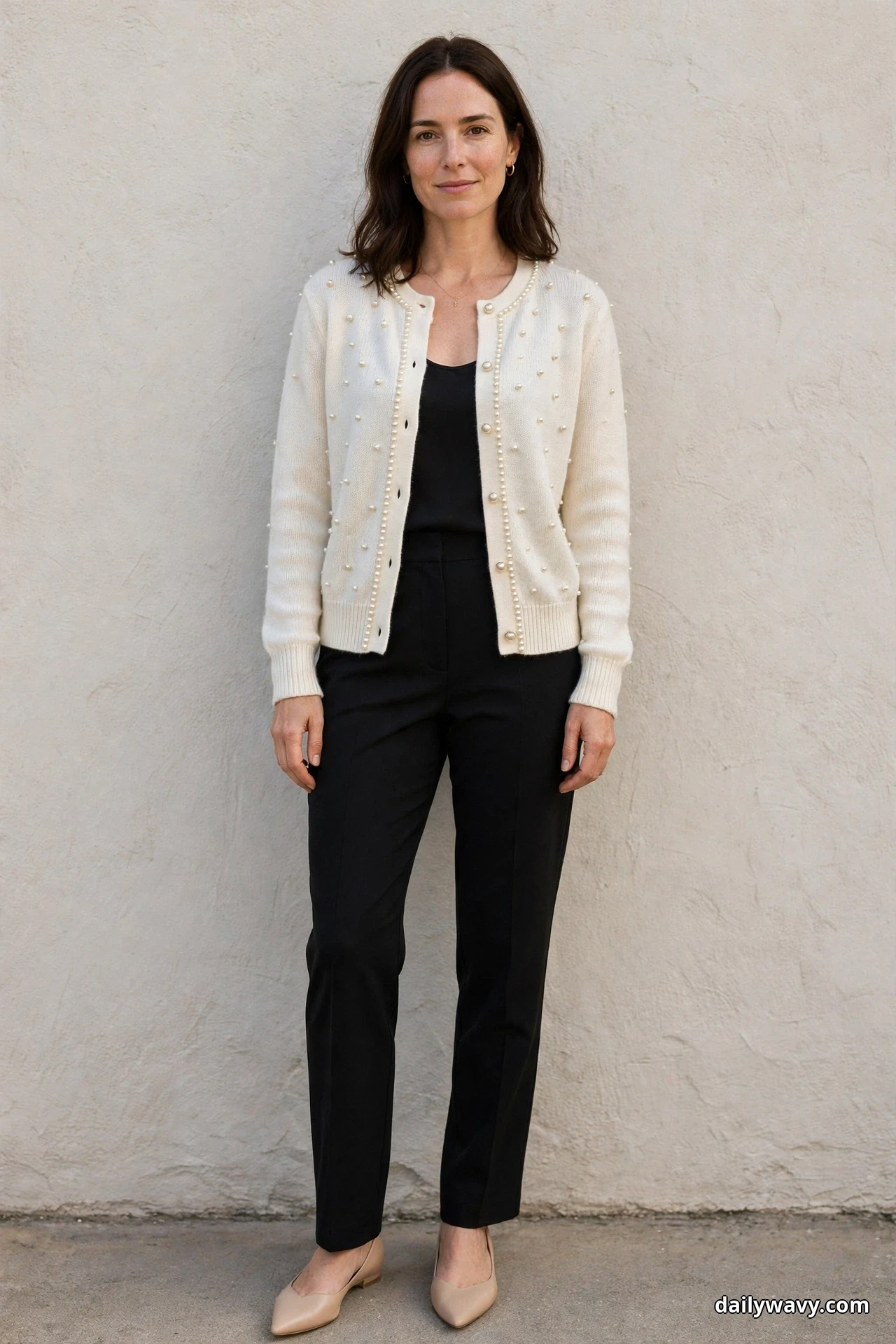 A woman stands full length against a textured cream wall wearing an ivory pearl-embellished cardigan open over a black camisole with black straight trousers and nude pointed flats