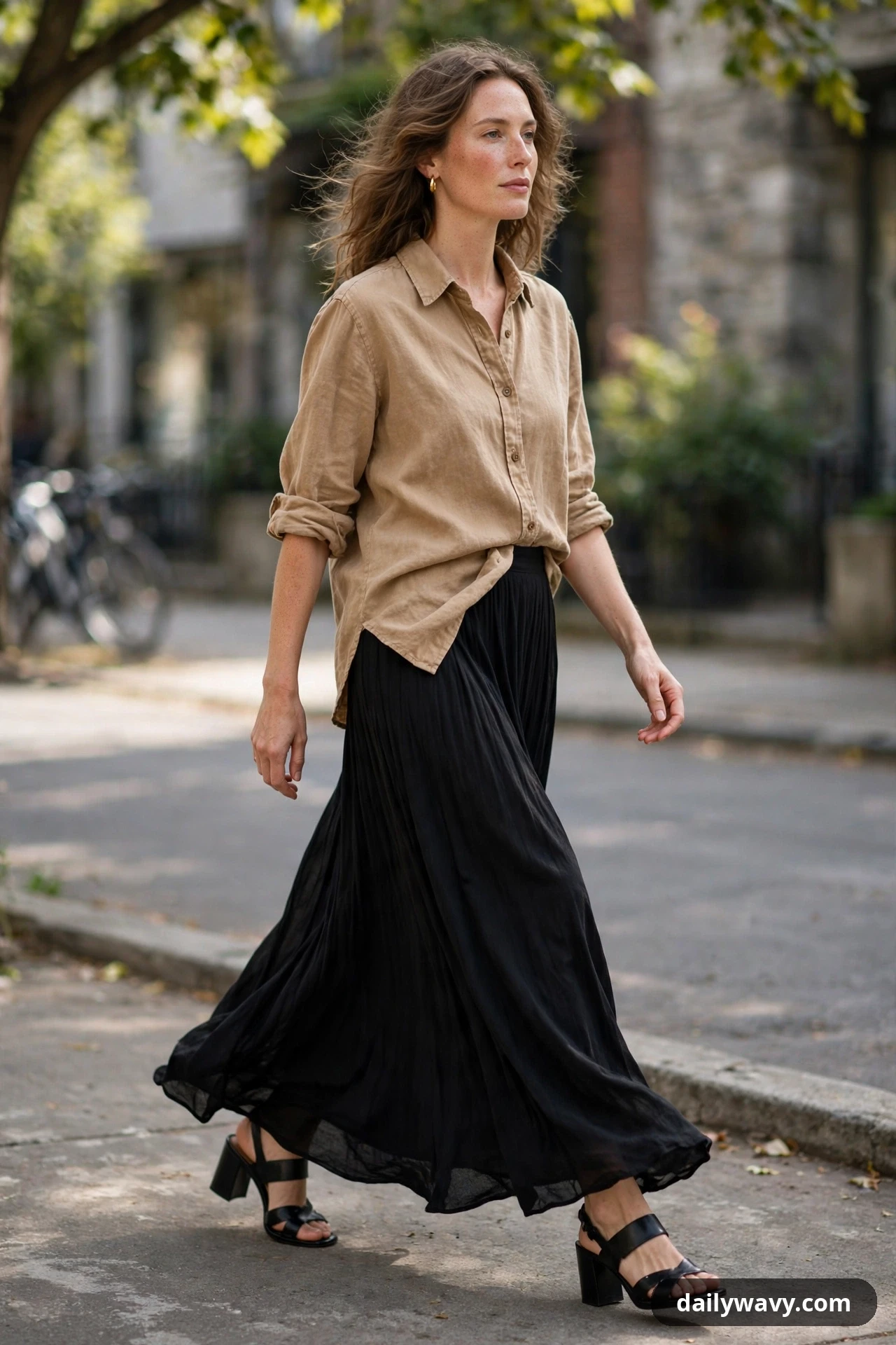 A woman wears a camel washed-linen button shirt half-tucked into a flowing black pleated maxi skirt with black block-heel sandals while walking outdoors.