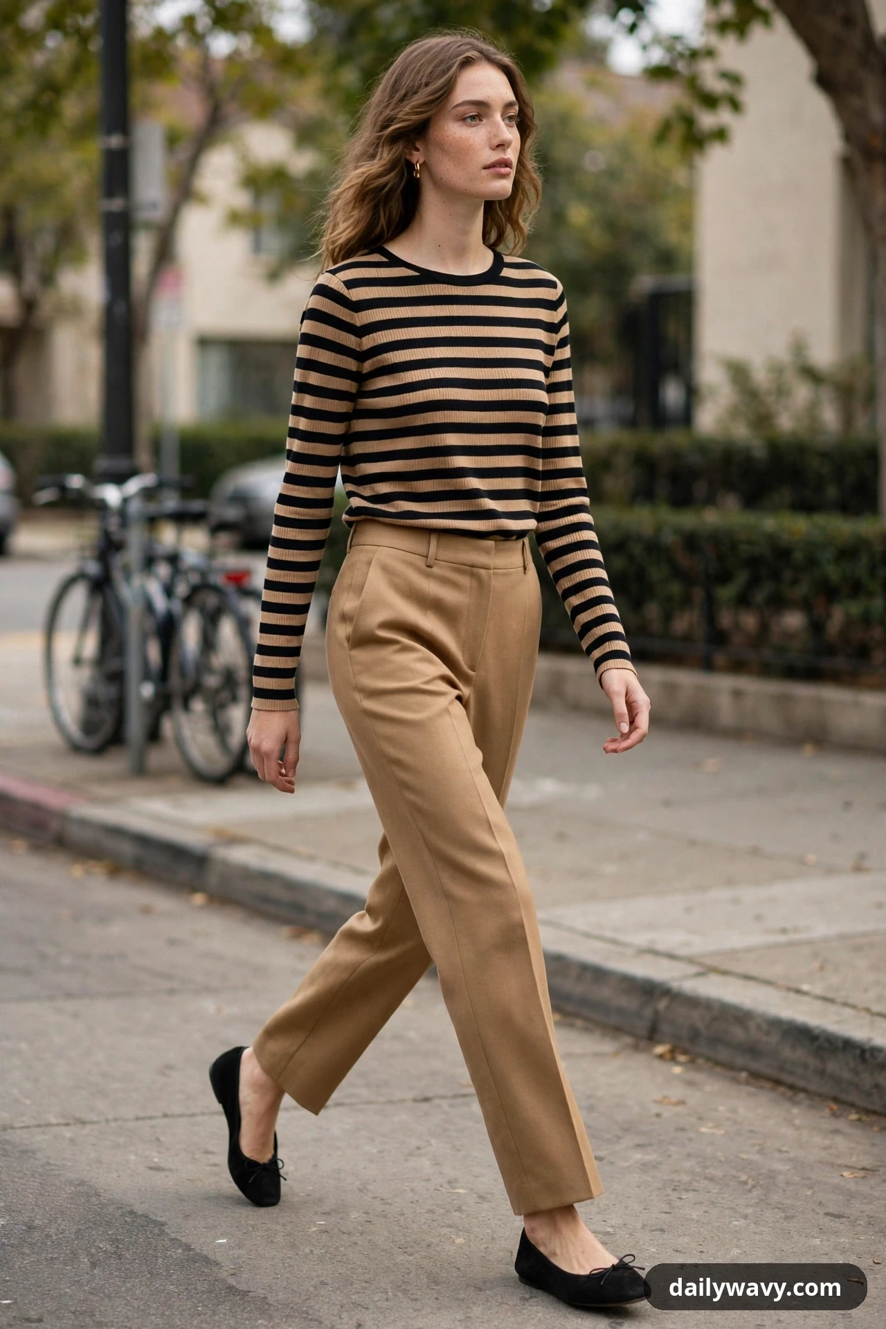 A woman wears a black-and-camel striped breton top tucked into solid camel straight-leg trousers with black ballet flats while crossing a street.