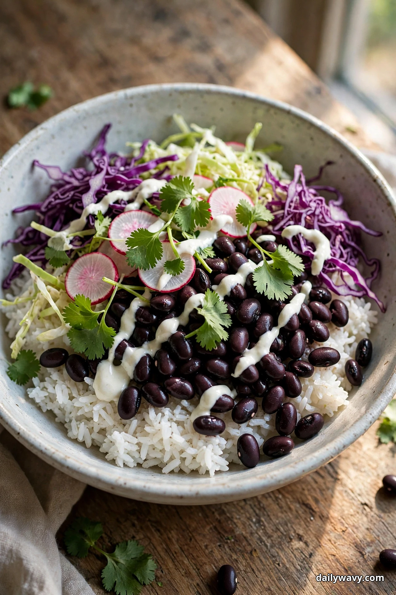 An overhead view of a speckled bowl of white rice topped with black beans, shredded cabbage, radish slices and cilantro leaves is drizzled with a white lime crema on a wooden table