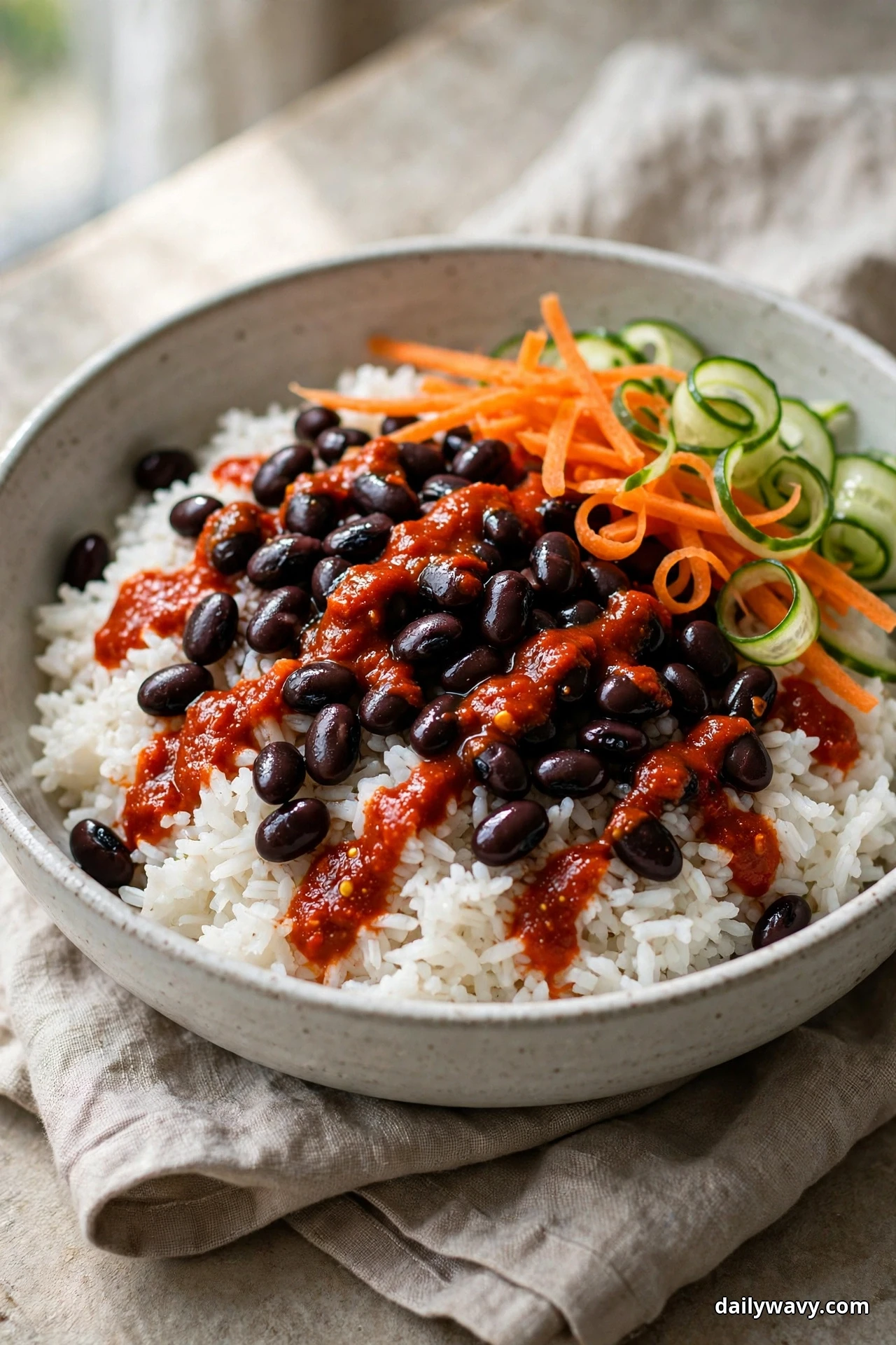 A speckled bowl of white rice topped with black beans and a red harissa drizzle sits beside carrot matchsticks and curled cucumber ribbons on linen