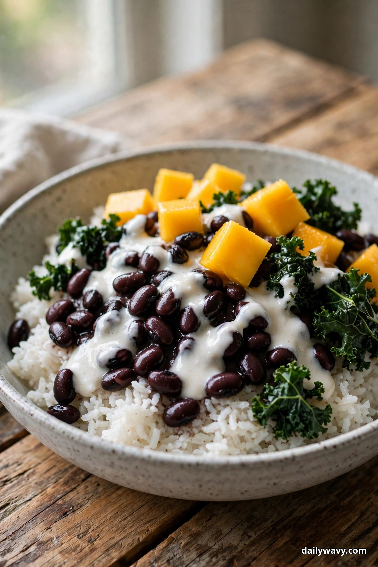 A speckled bowl of white rice topped with black beans under a creamy white coconut sauce is finished with kale leaves and mango cubes on a wooden table