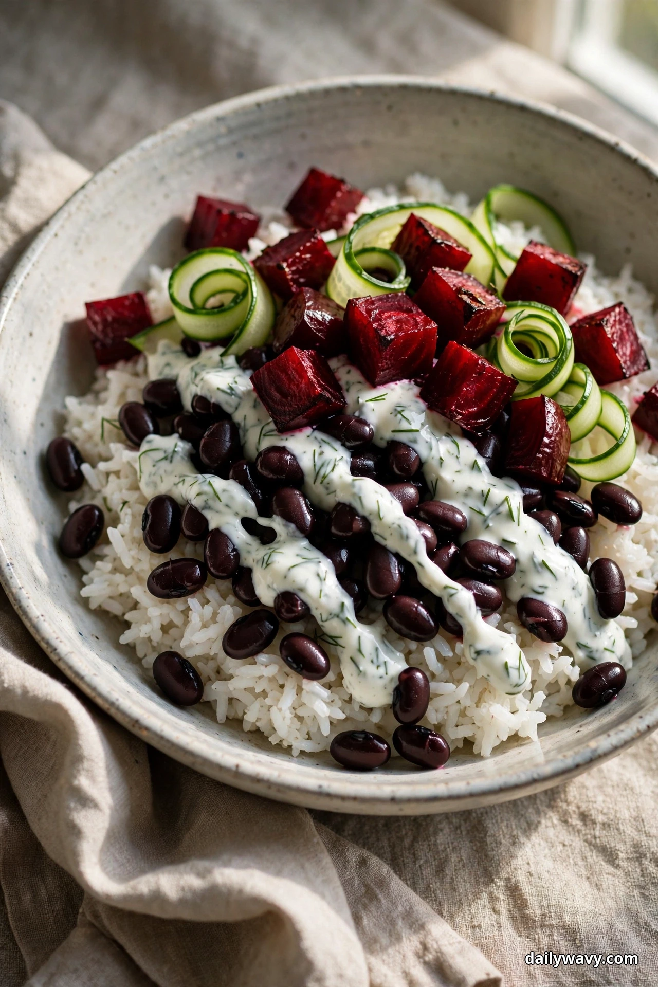 A speckled bowl of white rice topped with black beans and a dill-flecked yogurt sauce is finished with roasted beet cubes and curled cucumber ribbons over linen