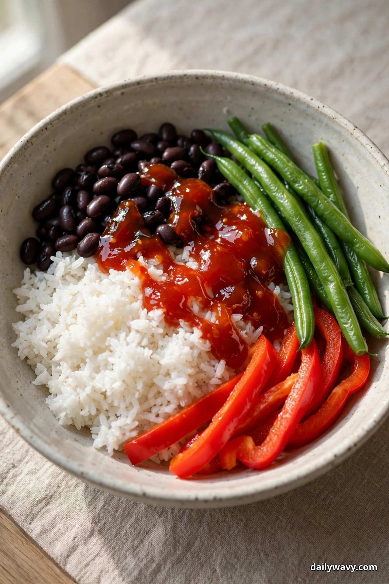 A speckled bowl of white rice topped with black beans and a glossy red sweet chili sauce sits beside whole green beans and red bell pepper strips on linen