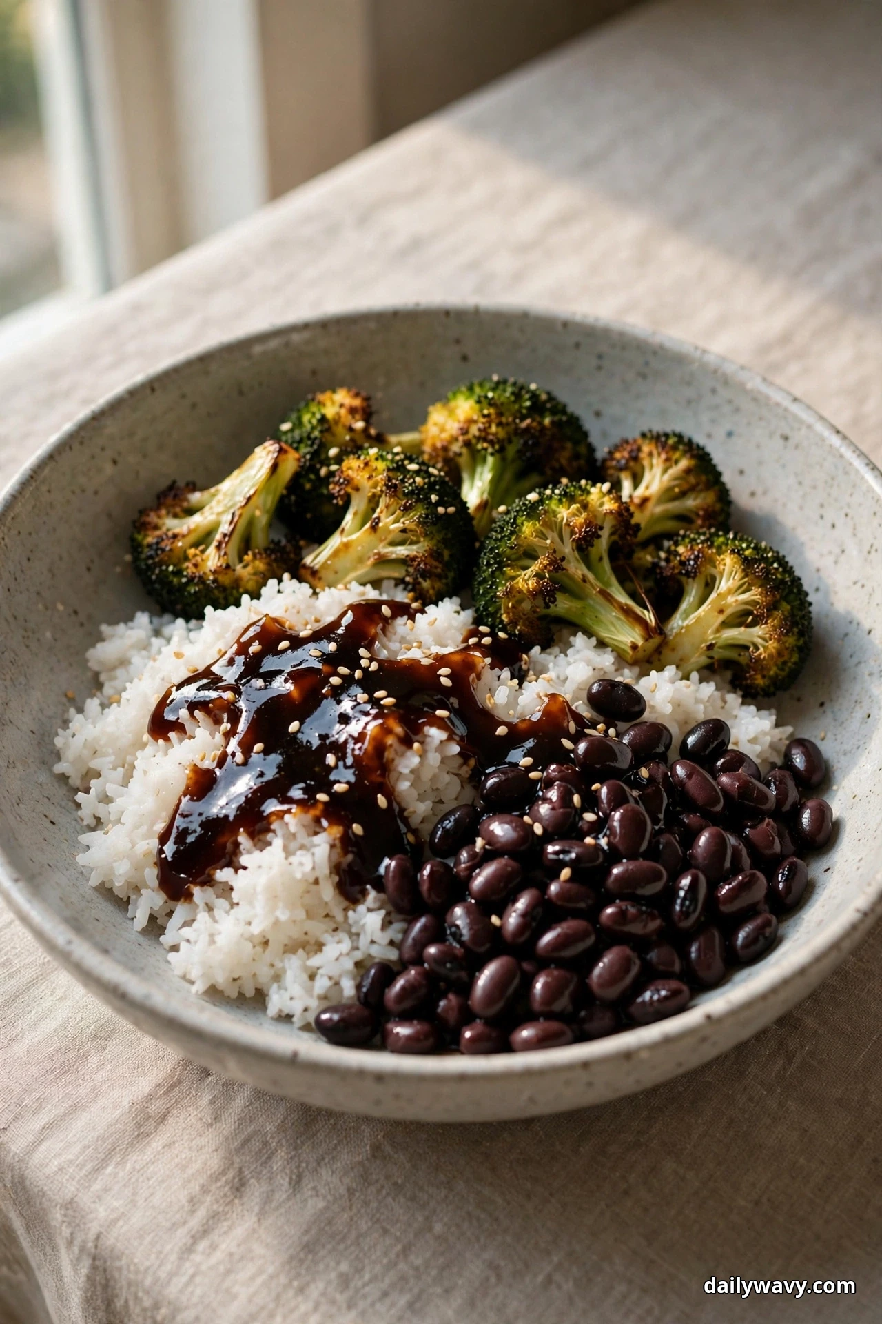 A speckled bowl of white rice topped with black beans, a dark teriyaki glaze and sesame seeds sits beside charred roasted broccoli florets on linen