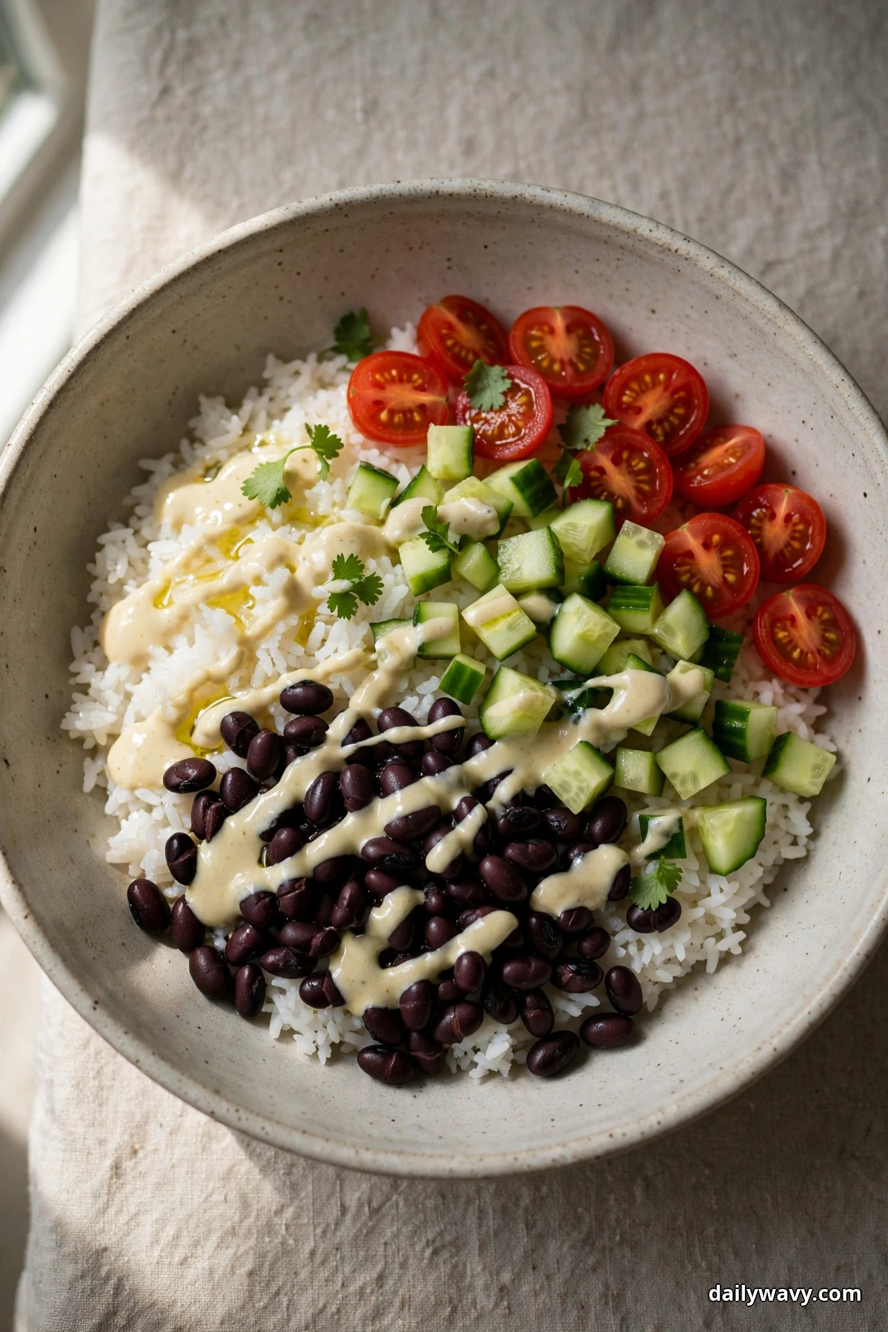 An overhead view of a speckled bowl of white rice topped with black beans and a pale lemon tahini drizzle sits beside diced cucumber and halved cherry tomatoes on linen