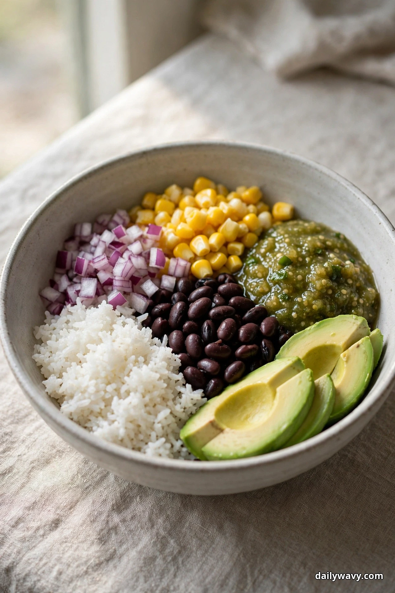 A speckled bowl holds sections of white rice, black beans, corn kernels, diced red onion and green salsa verde with fanned avocado slices, resting on linen