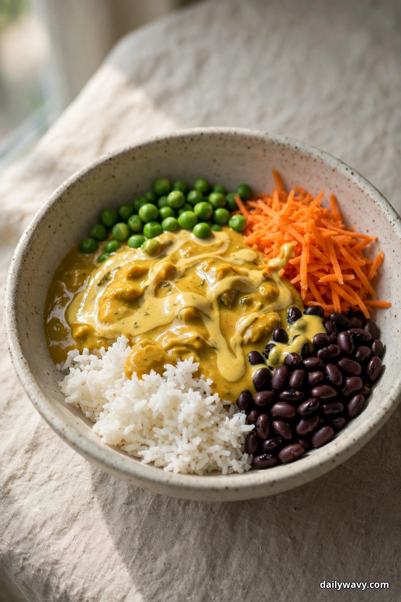 A speckled bowl of white rice and black beans covered with a golden curry yogurt sauce sits beside green peas and shredded carrot on linen