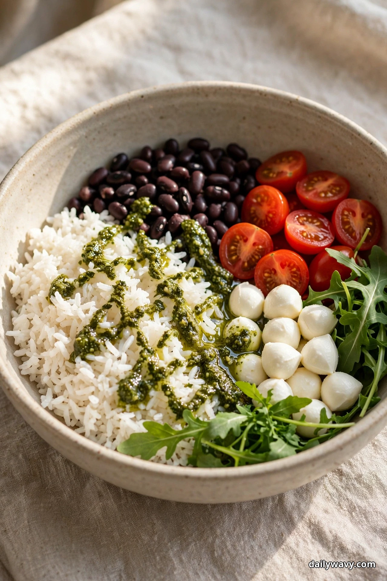 A speckled bowl of white rice drizzled with green pesto sits beside black beans, halved cherry tomatoes, mozzarella pearls and arugula leaves on linen