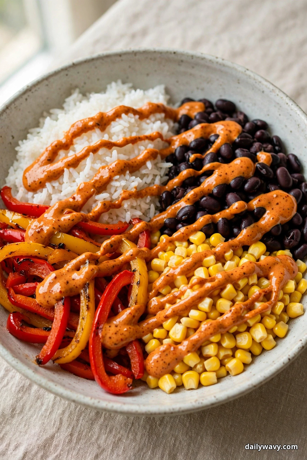 A speckled bowl of white rice and black beans drizzled with an orange chipotle crema sits beside sauteed bell pepper strips and corn kernels on linen