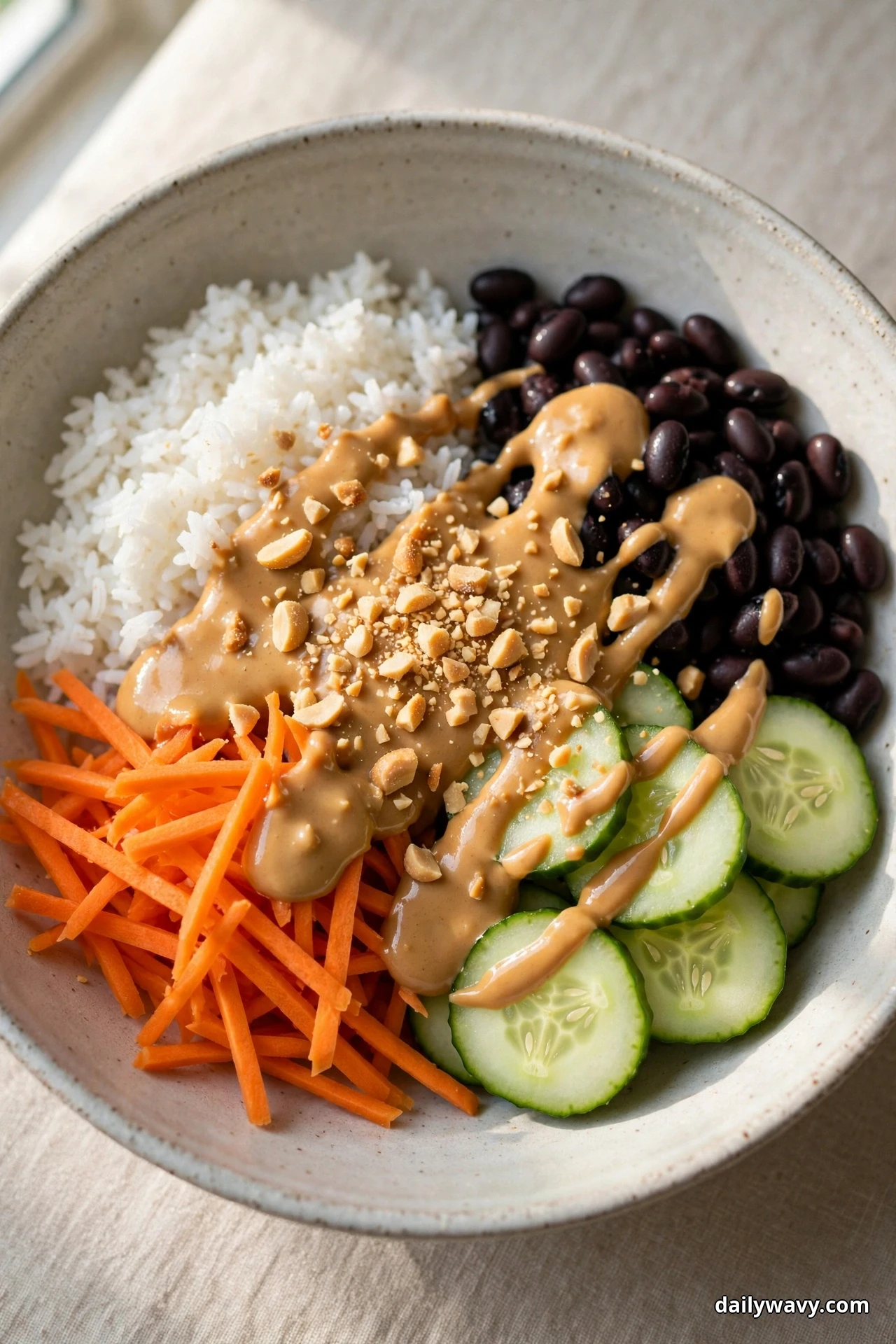 A speckled bowl of white rice and black beans covered with a creamy peanut sauce and crushed peanuts sits beside carrot matchsticks and cucumber slices on linen