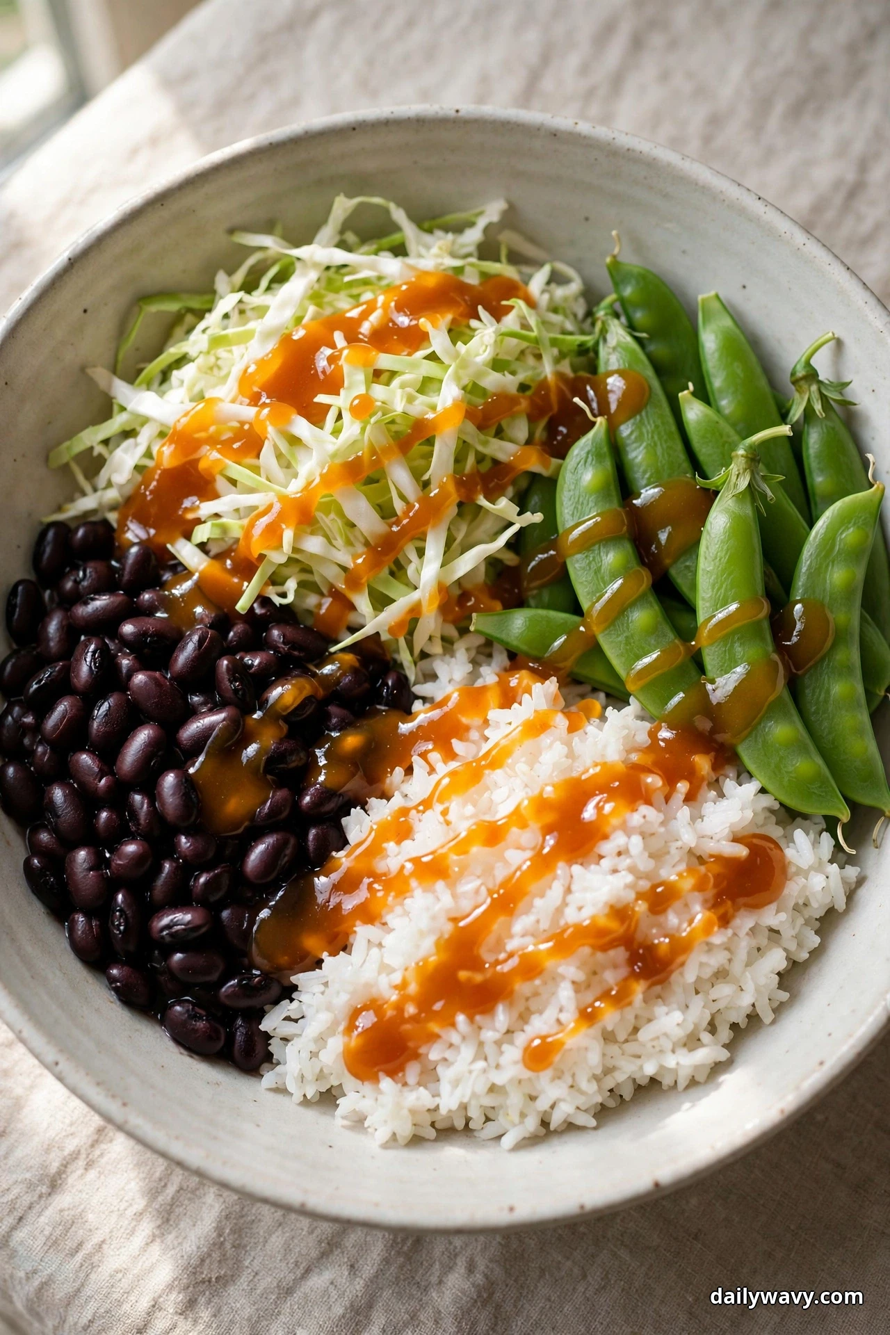 A speckled bowl of white rice and black beans drizzled with a glossy amber orange glaze sits beside shredded cabbage and whole sugar snap peas on linen