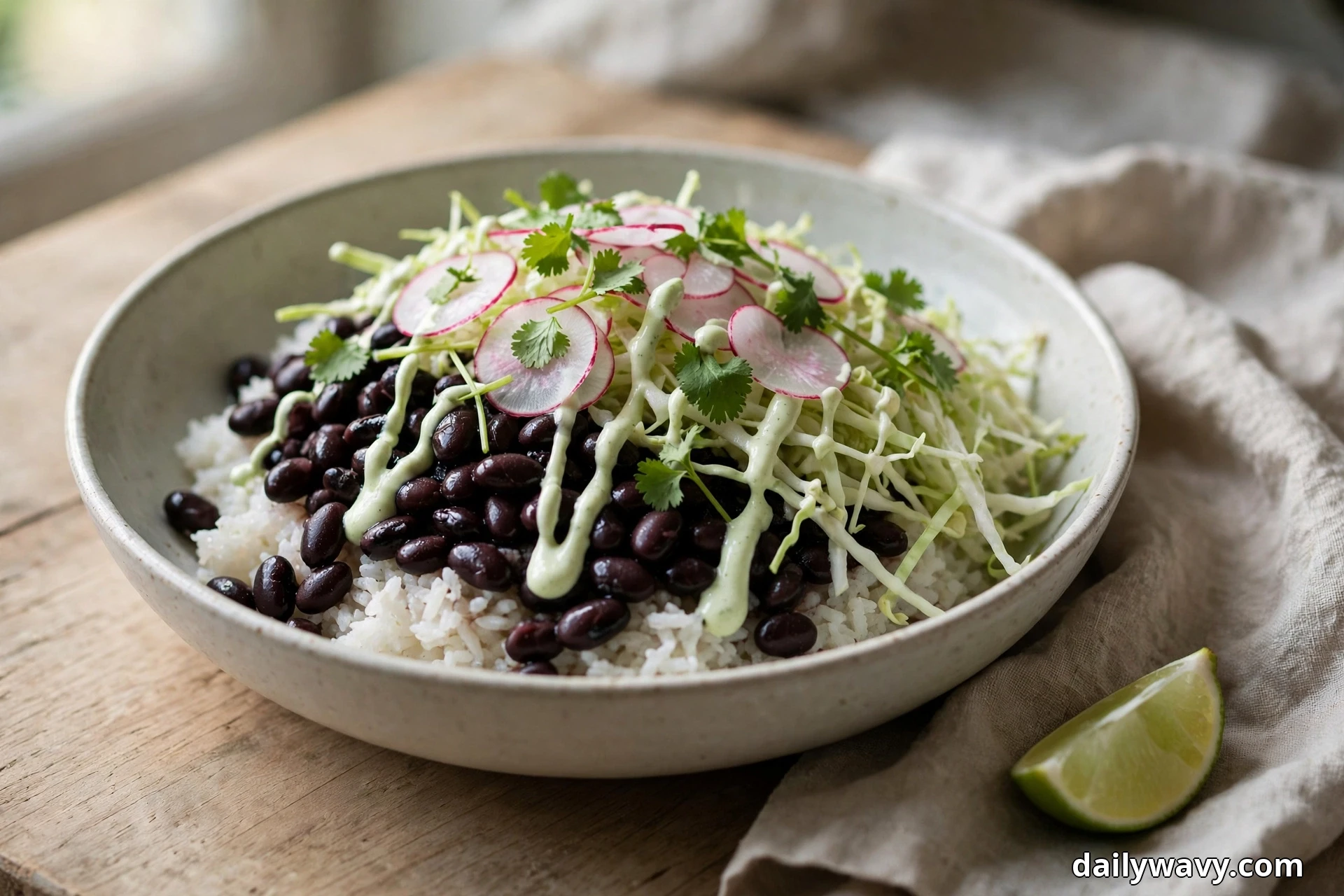 A shallow ceramic bowl of white rice topped with black beans, shredded cabbage, radish slices and cilantro leaves is drizzled with a pale lime crema and sits on a wooden table beside a lime wedge