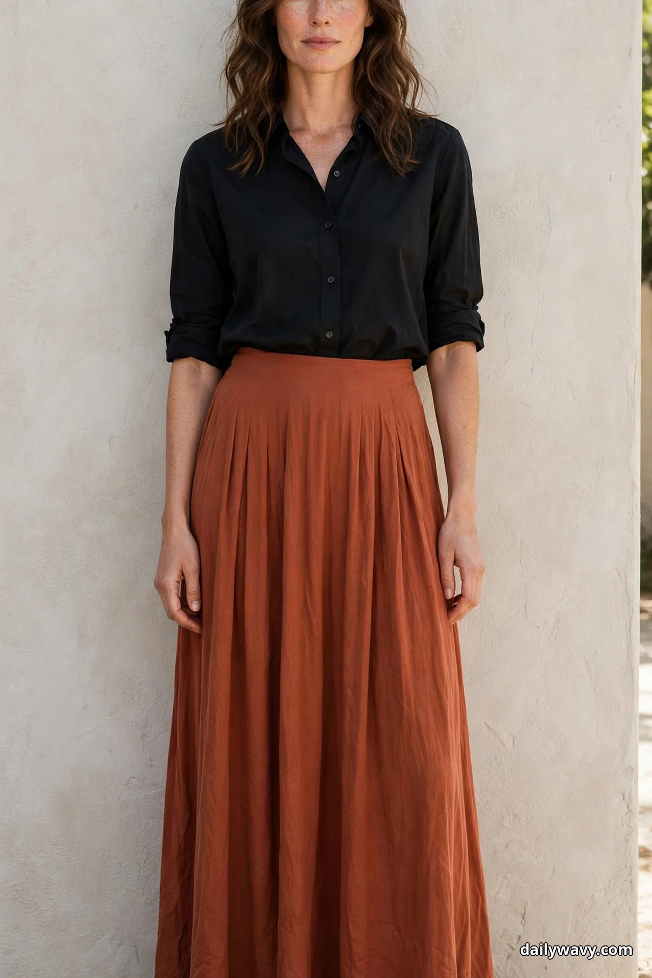 A woman wearing a black button-front blouse with rolled sleeves and a flowing terracotta maxi skirt stands against a light stucco wall, framed from the chin to the hem of the skirt