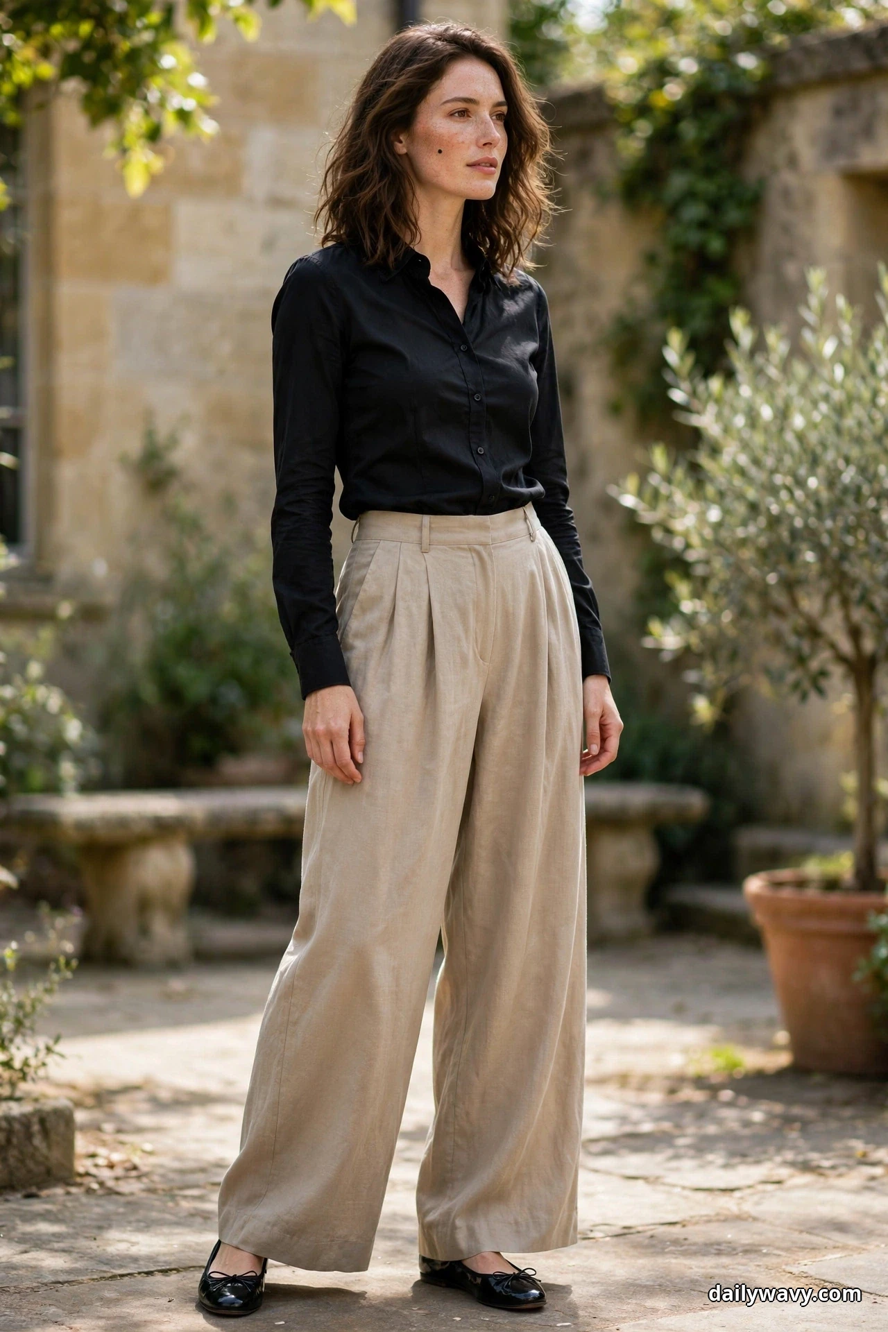 A woman wearing a fitted black button-front blouse tucked into flowing stone wide-leg trousers with black ballet flats stands in a garden courtyard, shown full length