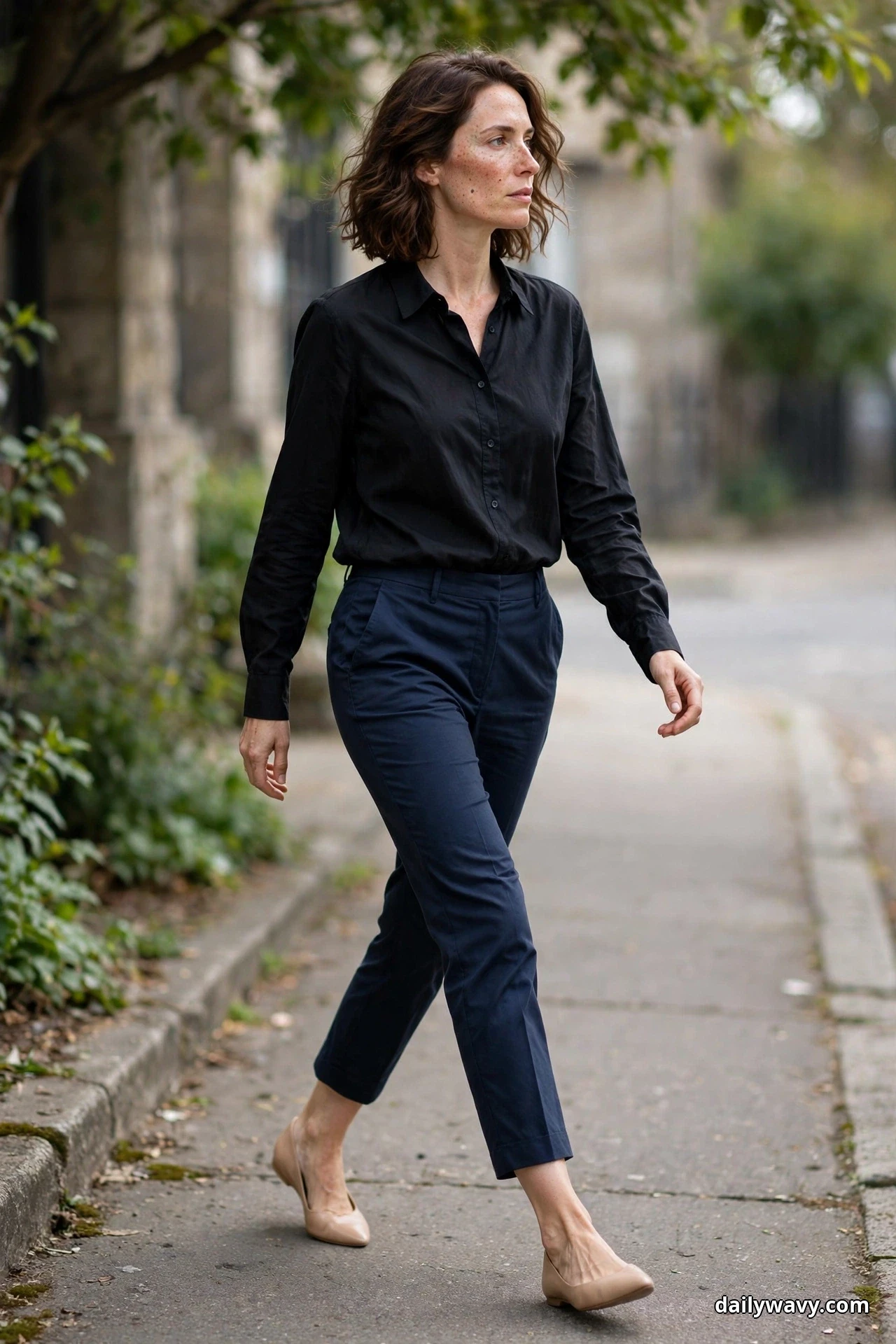 A woman walks along a sidewalk wearing a black blouse tucked into navy cropped ankle trousers with nude pointed flats, shown full length