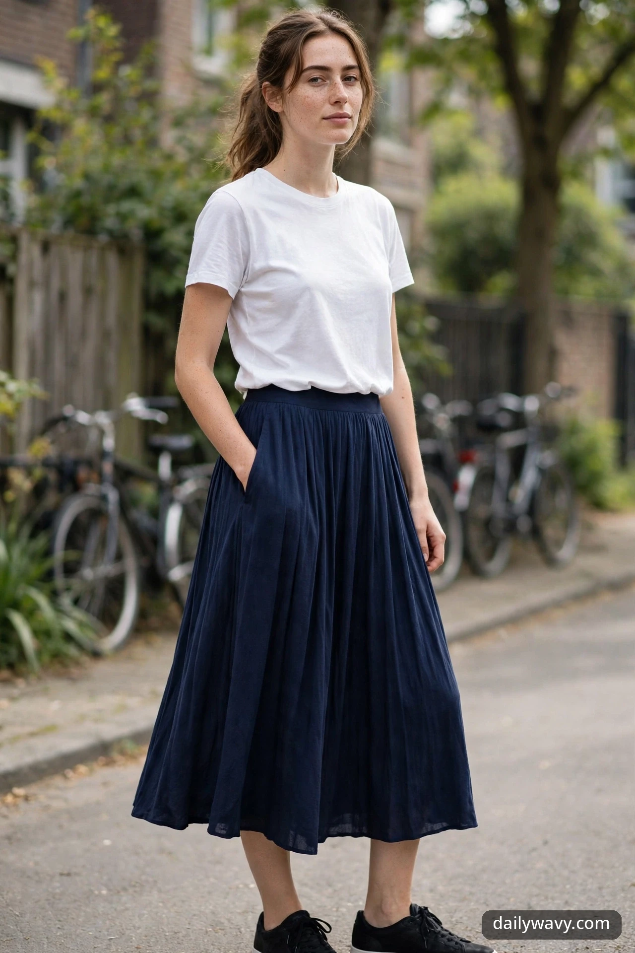 A woman walking on a street with parked bicycles in a white crewneck tee, a navy pleated midi skirt and black sneakers.