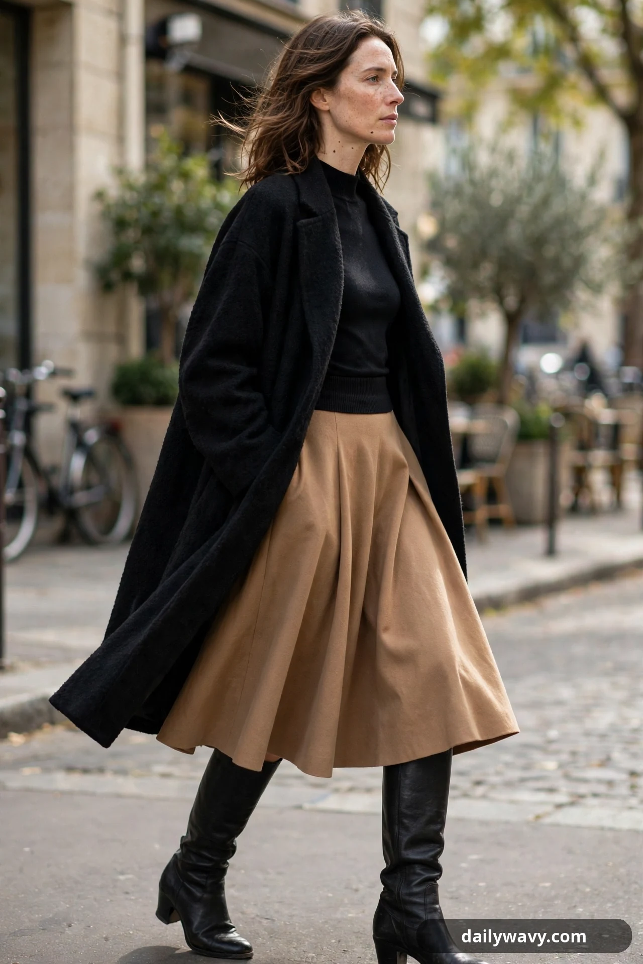A woman walks along a city street in a long open black wool coat over a fitted black mock-neck top, a flared camel A-line skirt and black knee-high boots.
