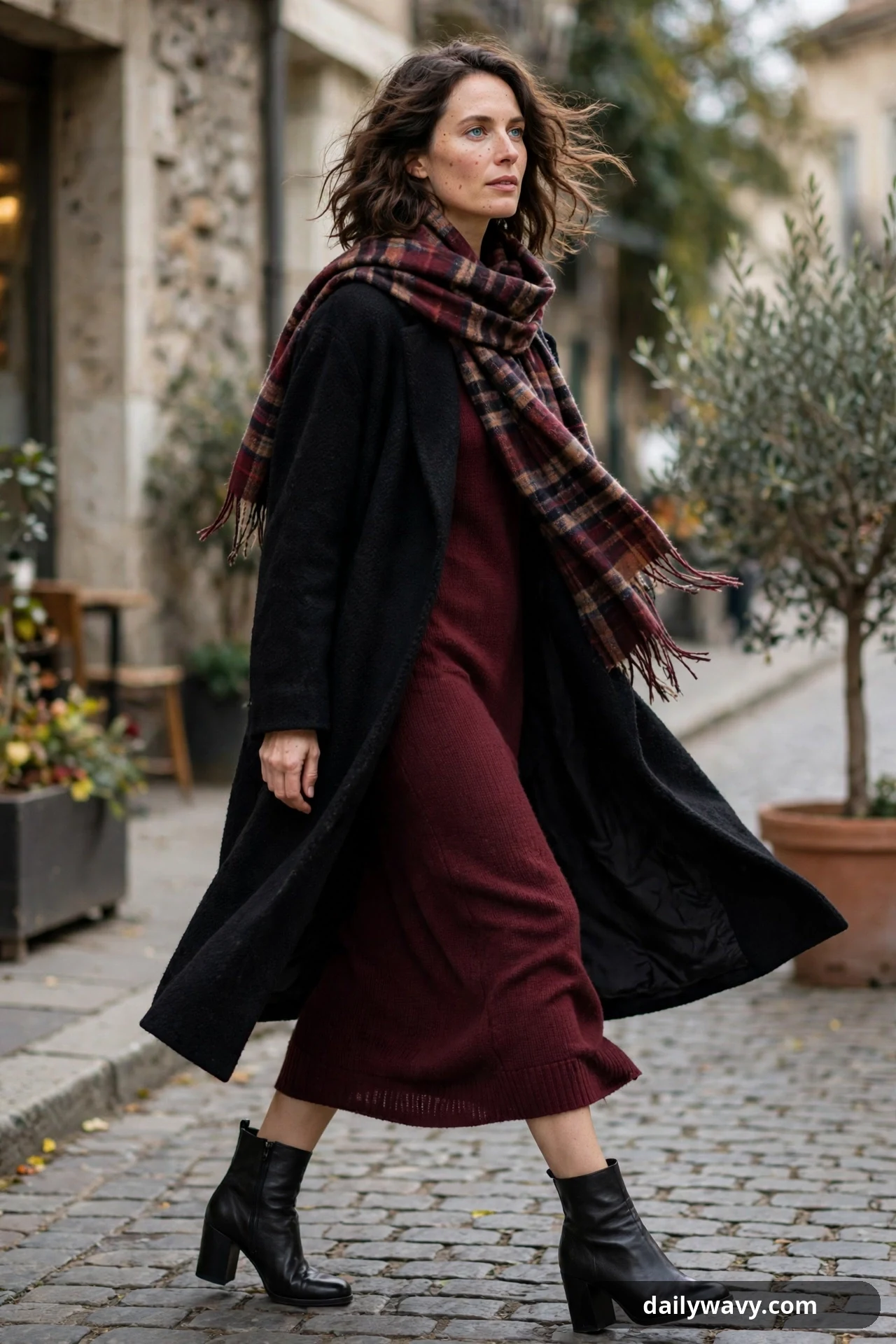 A woman walks across a cobbled street in a long open black wool coat over a burgundy rib-knit midi dress, with a burgundy plaid scarf at her neck and black block-heel ankle boots.