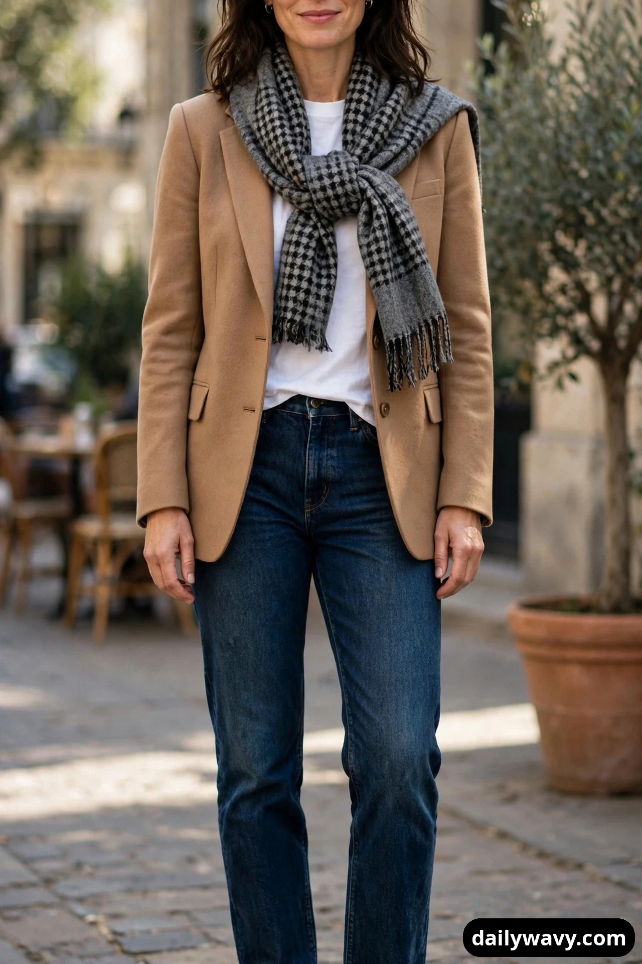 A woman wears a white crewneck tee under a camel blazer with dark indigo jeans, and a grey-and-black houndstooth blanket scarf knotted loosely at her chest.