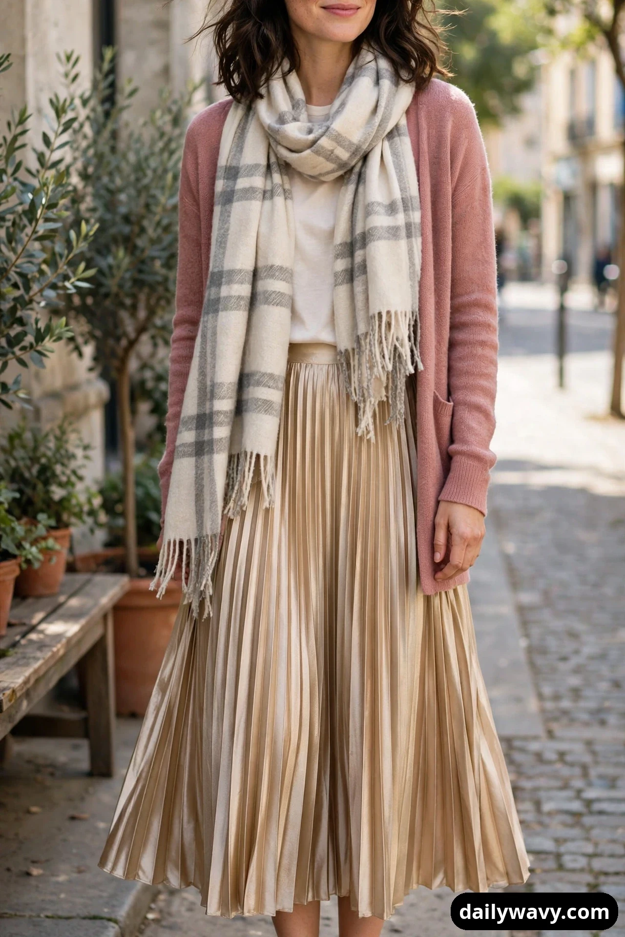 A woman wears a dusty-rose open cardigan over a cream top with a pleated champagne midi skirt, and a cream-and-grey plaid blanket scarf looped around her neck.