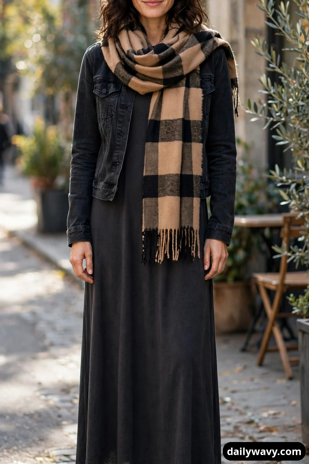 A woman wears a charcoal maxi dress under a cropped black denim jacket, with a camel-and-black checked blanket scarf wrapped around her neck.